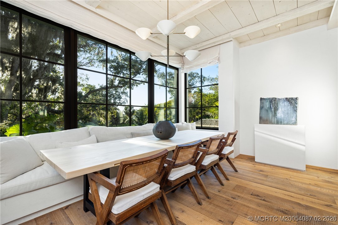 Dining area with a long white table, wood chairs, banquette seating, and floor-to-ceiling garden-view windows.