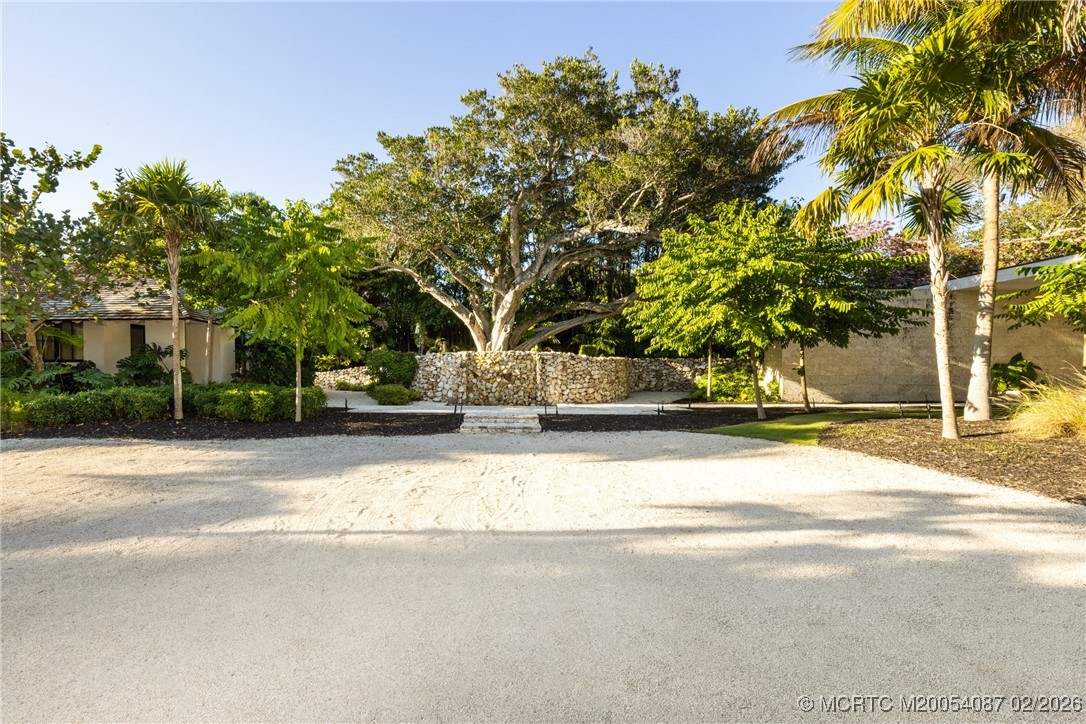 Gravel driveway and landscaped entrance framed by a stone wall, mature tree, palms, and partial buildings.