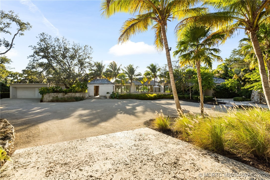 Single-story home behind a broad driveway, framed by tall palms and tropical landscaping.