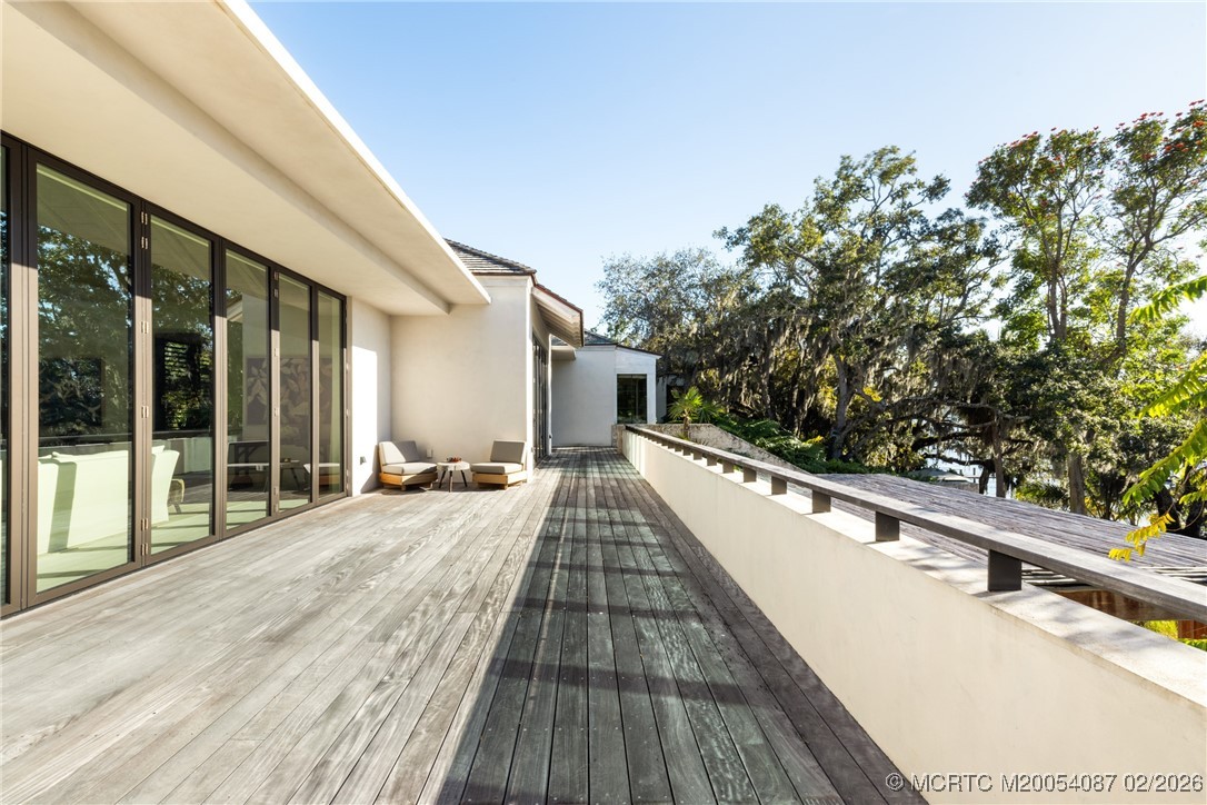 Wide wood deck beside a modern home, with glass doors, lounge chairs, and leafy trees beyond the railing.