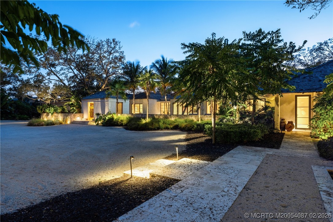 Single-story home with a broad gravel driveway, tropical landscaping, and warmly lit windows at dusk.