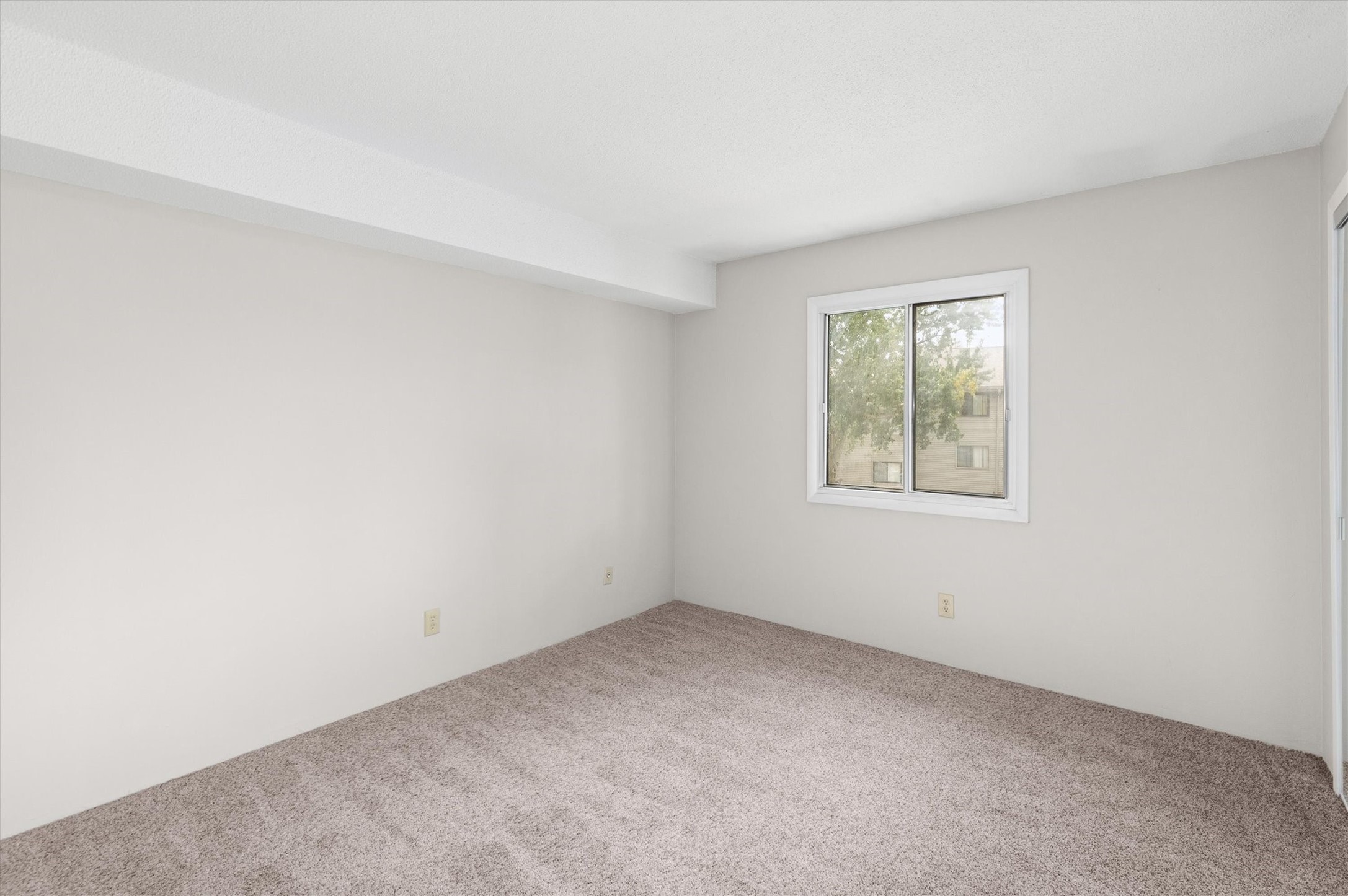 Empty carpeted room with neutral walls and a window overlooking a neighboring building.