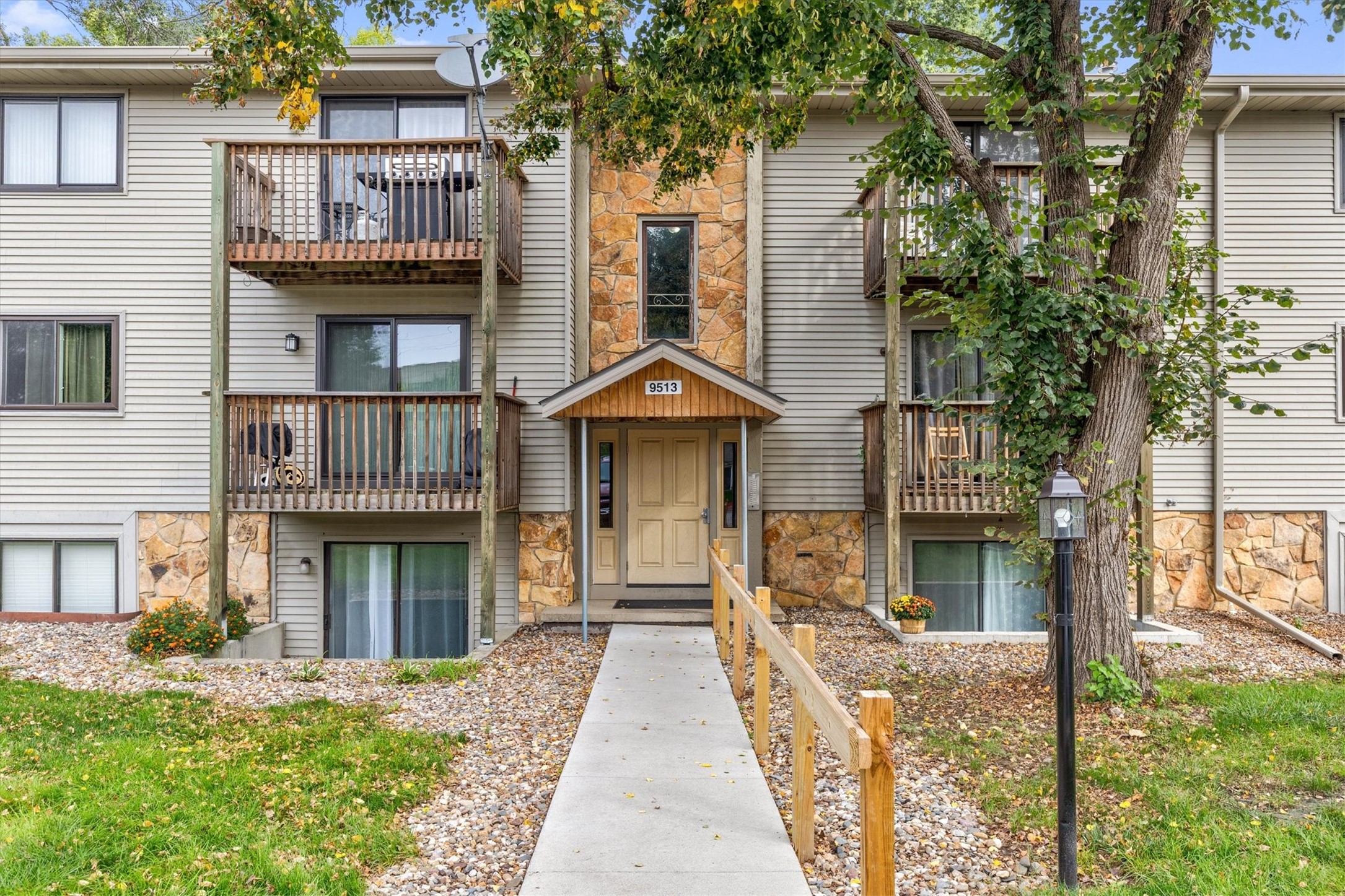 Residential building entrance with stone accents, wooden balconies, and a walkway beneath mature trees.