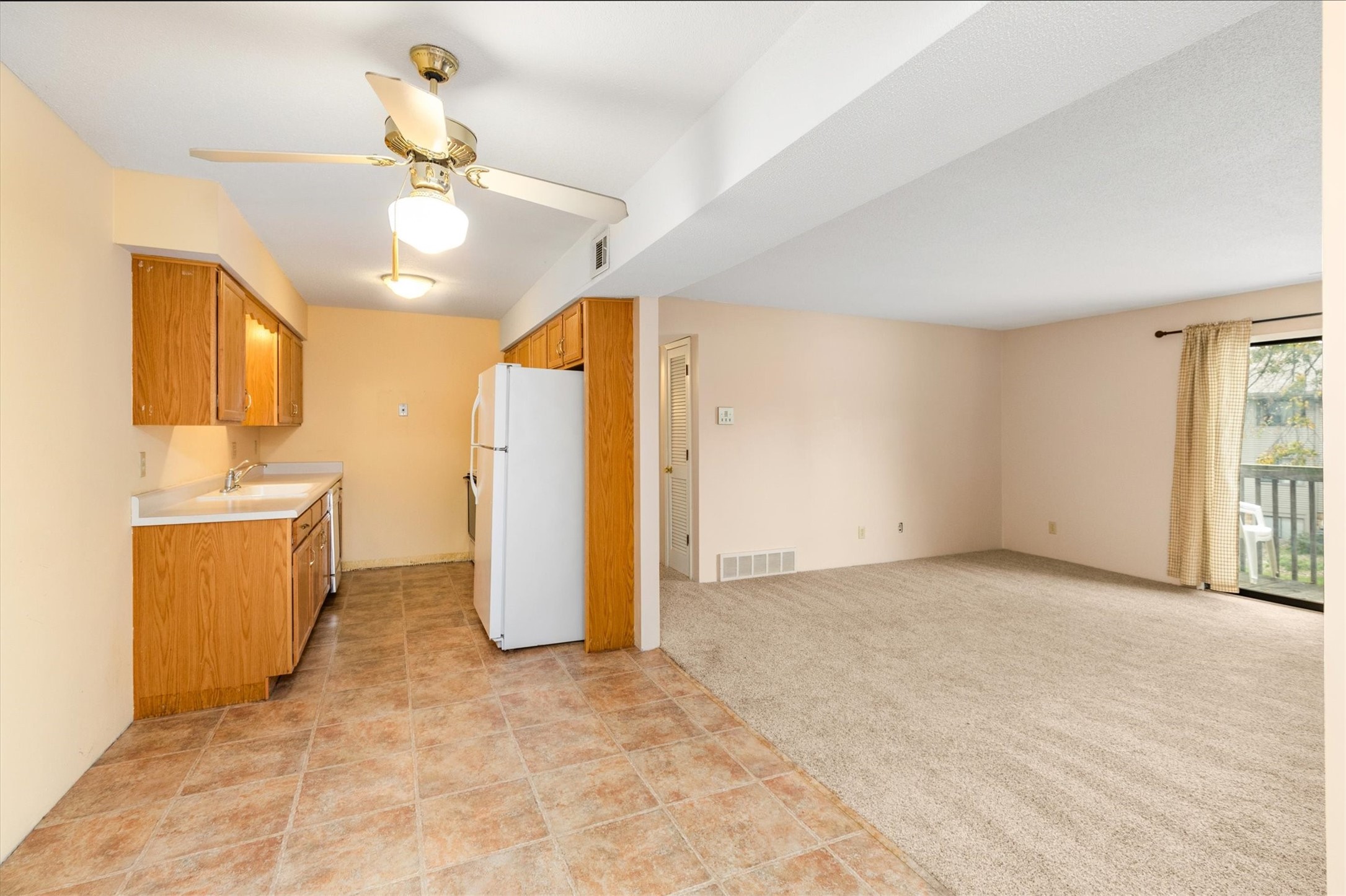 Empty kitchen and living room with oak cabinets, tiled flooring, beige carpet, and a sliding glass door.