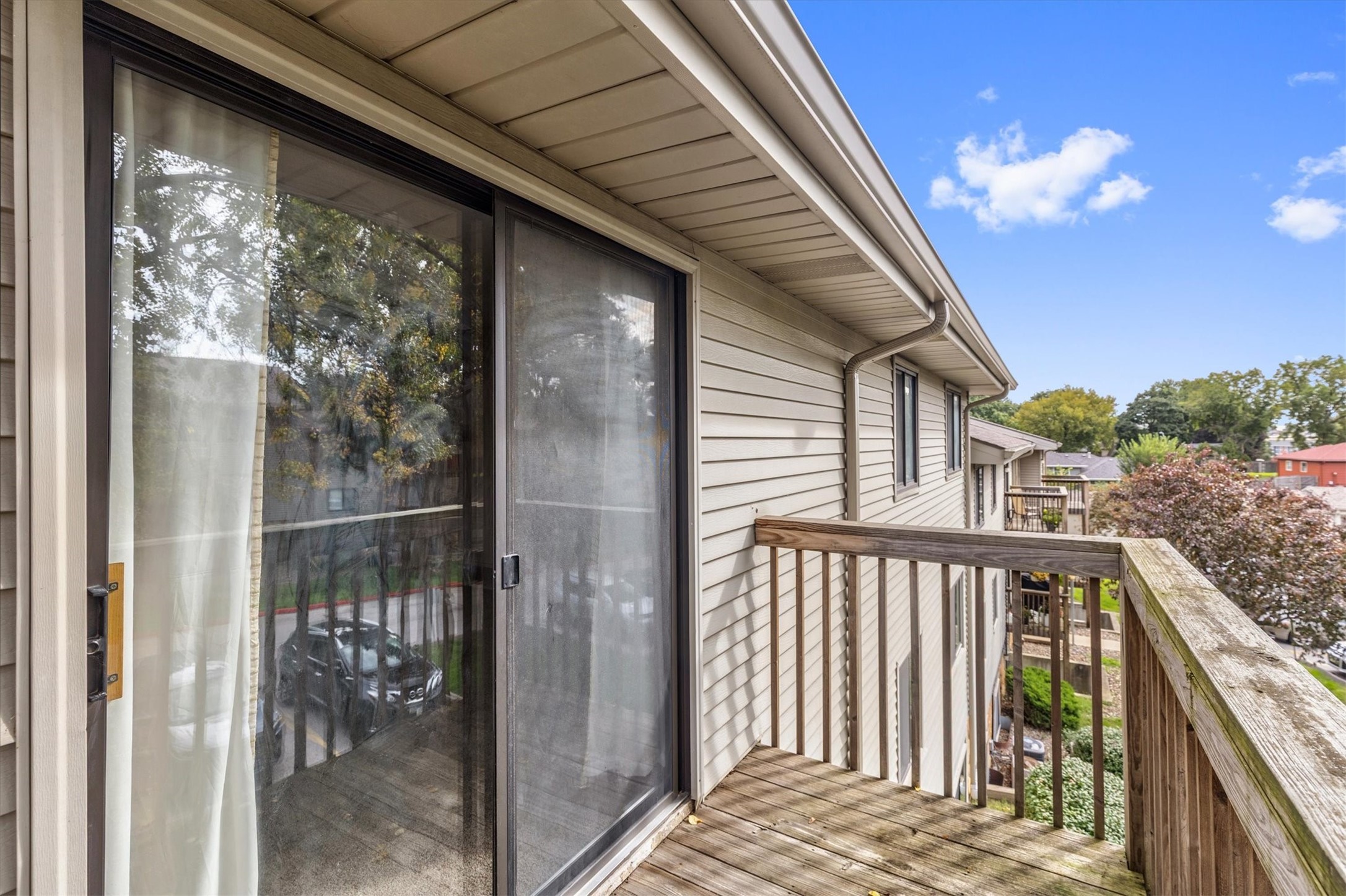Wooden balcony beside beige siding, with a large sliding glass door and neighboring homes beyond.