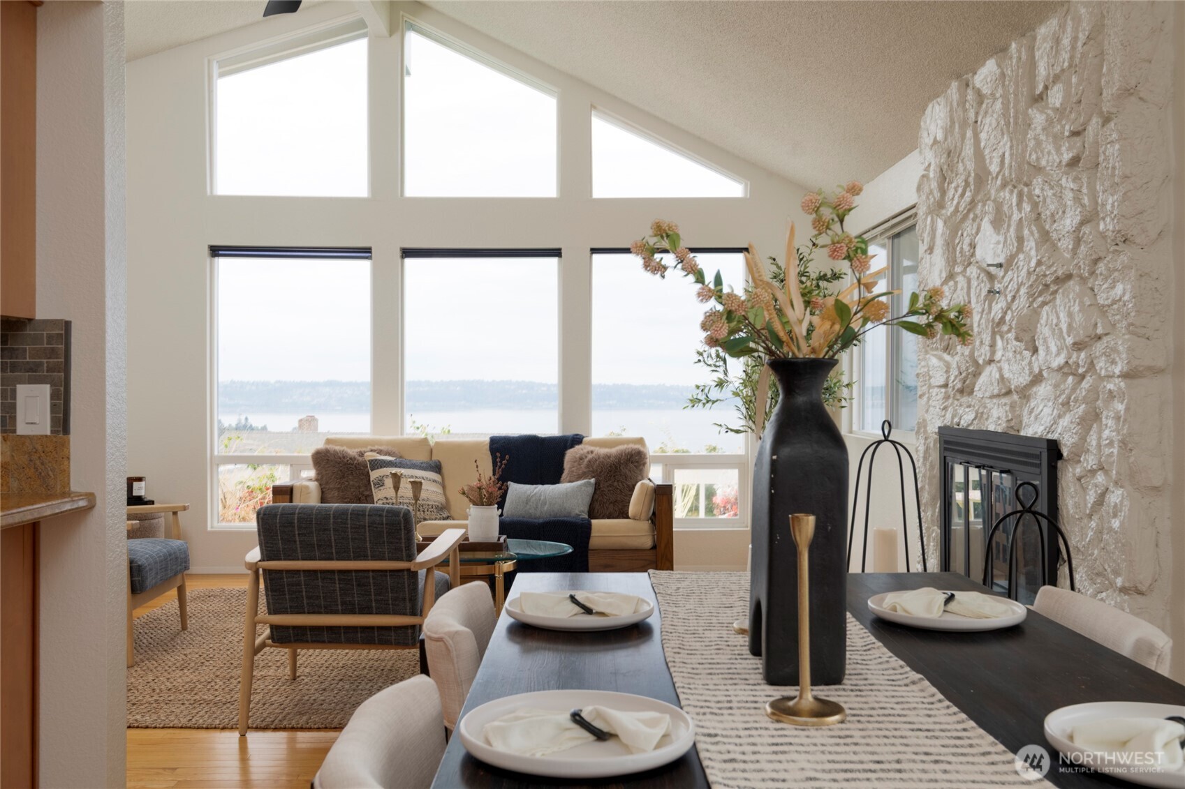 Living and dining room with a stone fireplace, vaulted ceiling, and water views through tall windows.