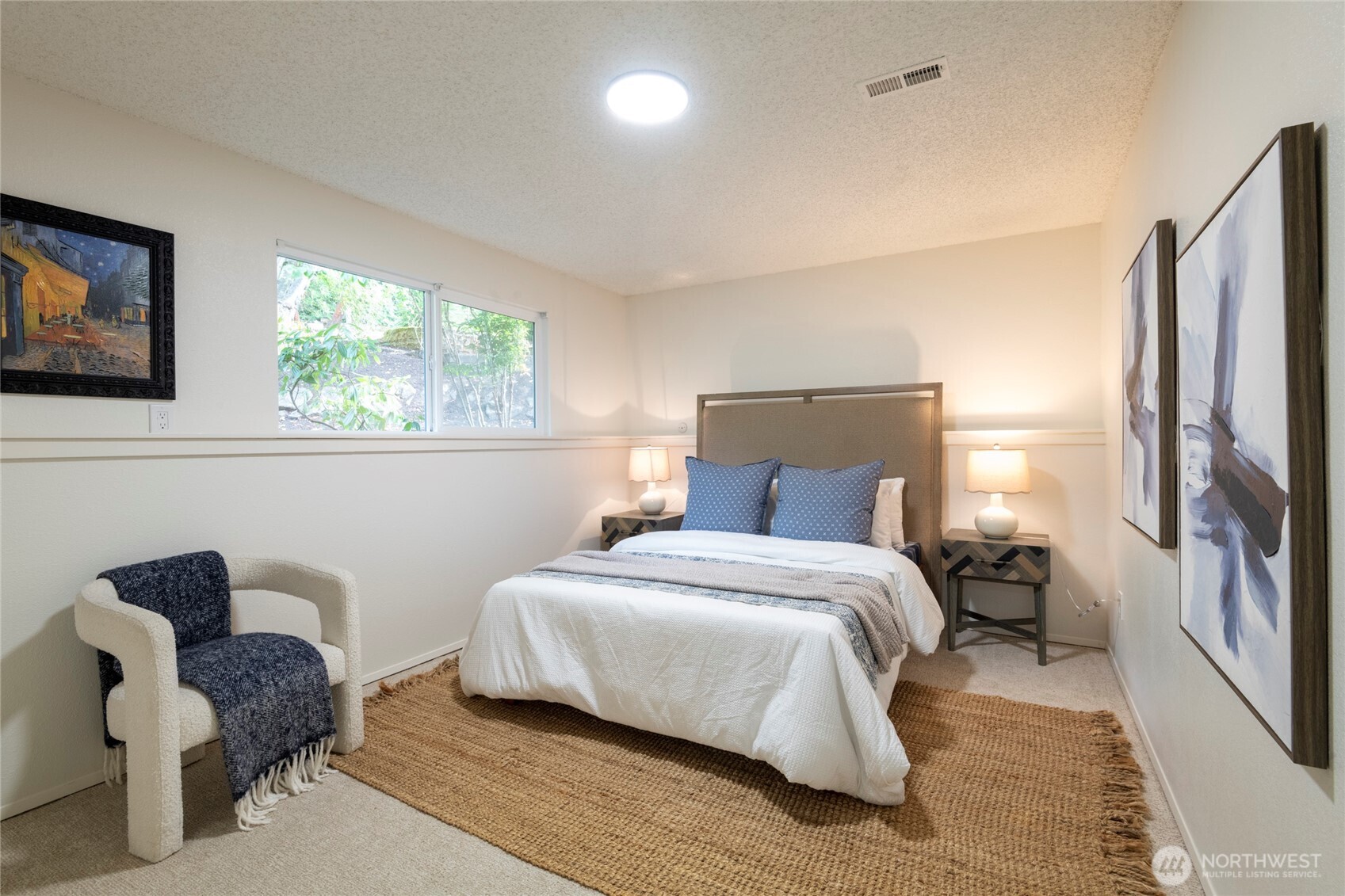 Staged bedroom with a white bed, blue pillows, woven rug, and window overlooking greenery.