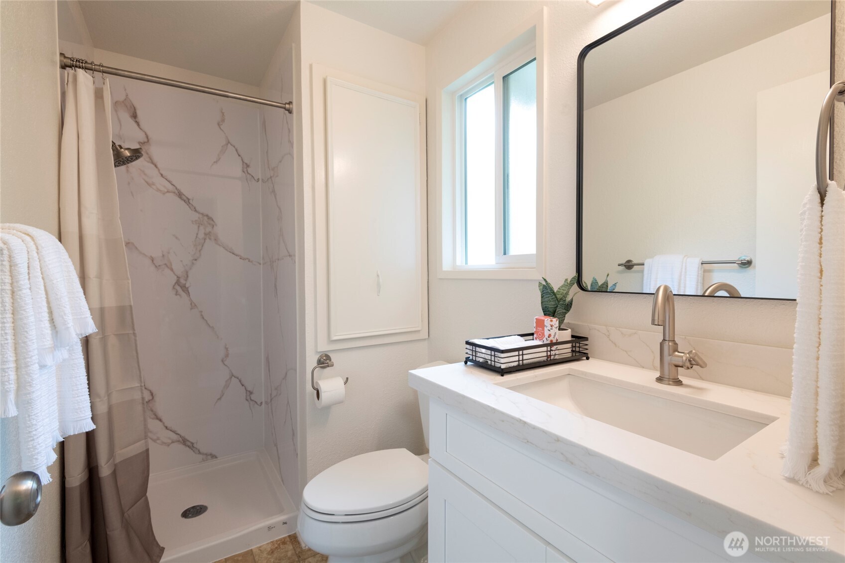 Bathroom with a marble-look shower surround, white vanity, black-framed mirror, and window.