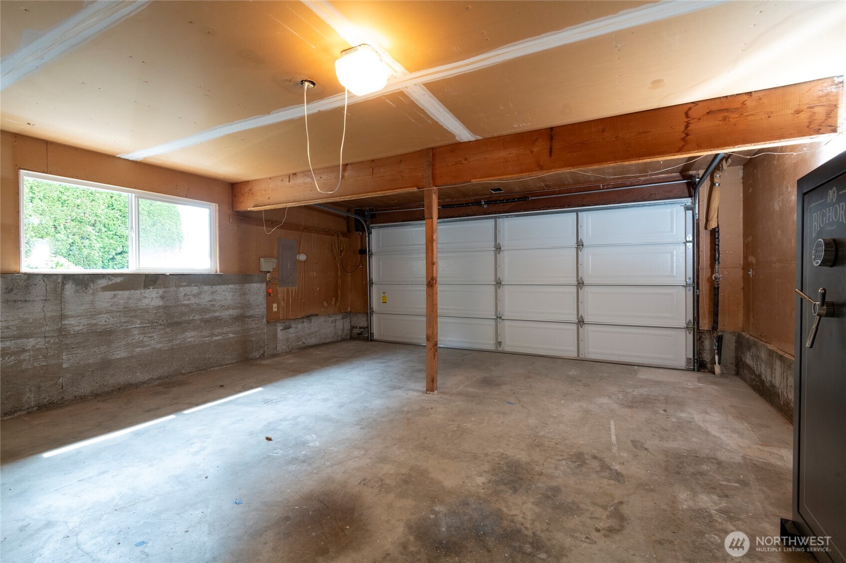 Empty garage with exposed beams, concrete floor, white garage door, and a large safe.