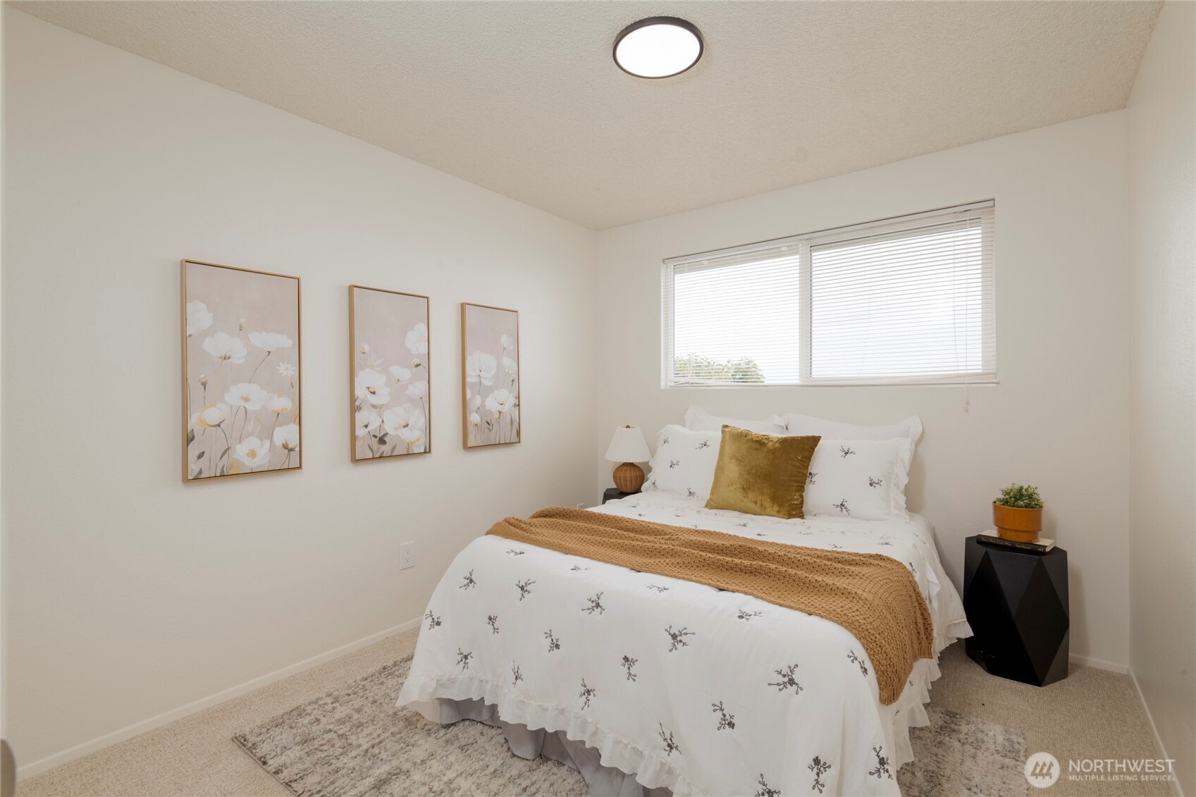 Staged bedroom with a white bed, mustard-gold accents, floral wall art, and a wide window.