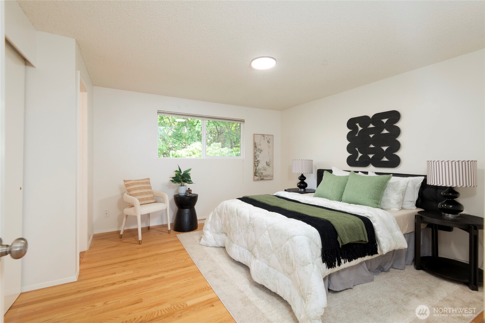 Furnished bedroom with a white bed, green accents, hardwood floors, and a window overlooking trees.