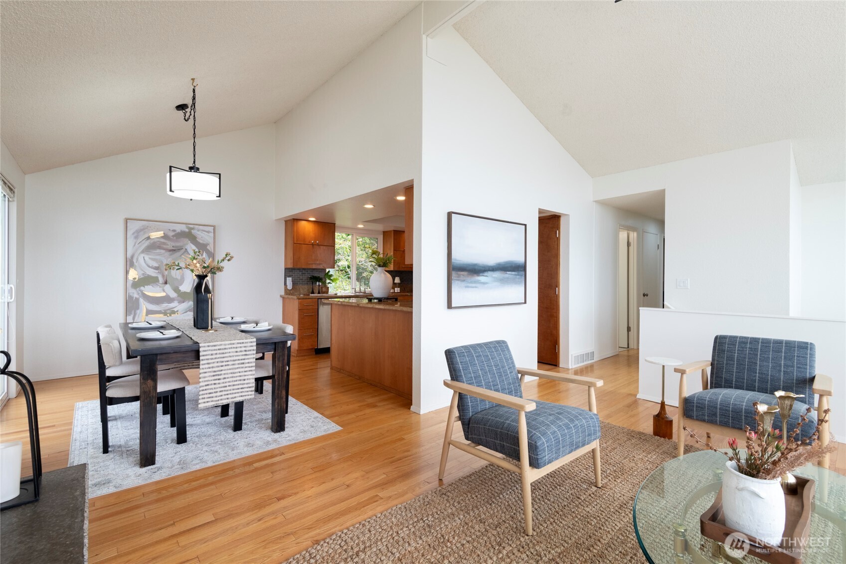 Open-plan living and dining room with vaulted ceiling, hardwood floors, and adjoining kitchen.