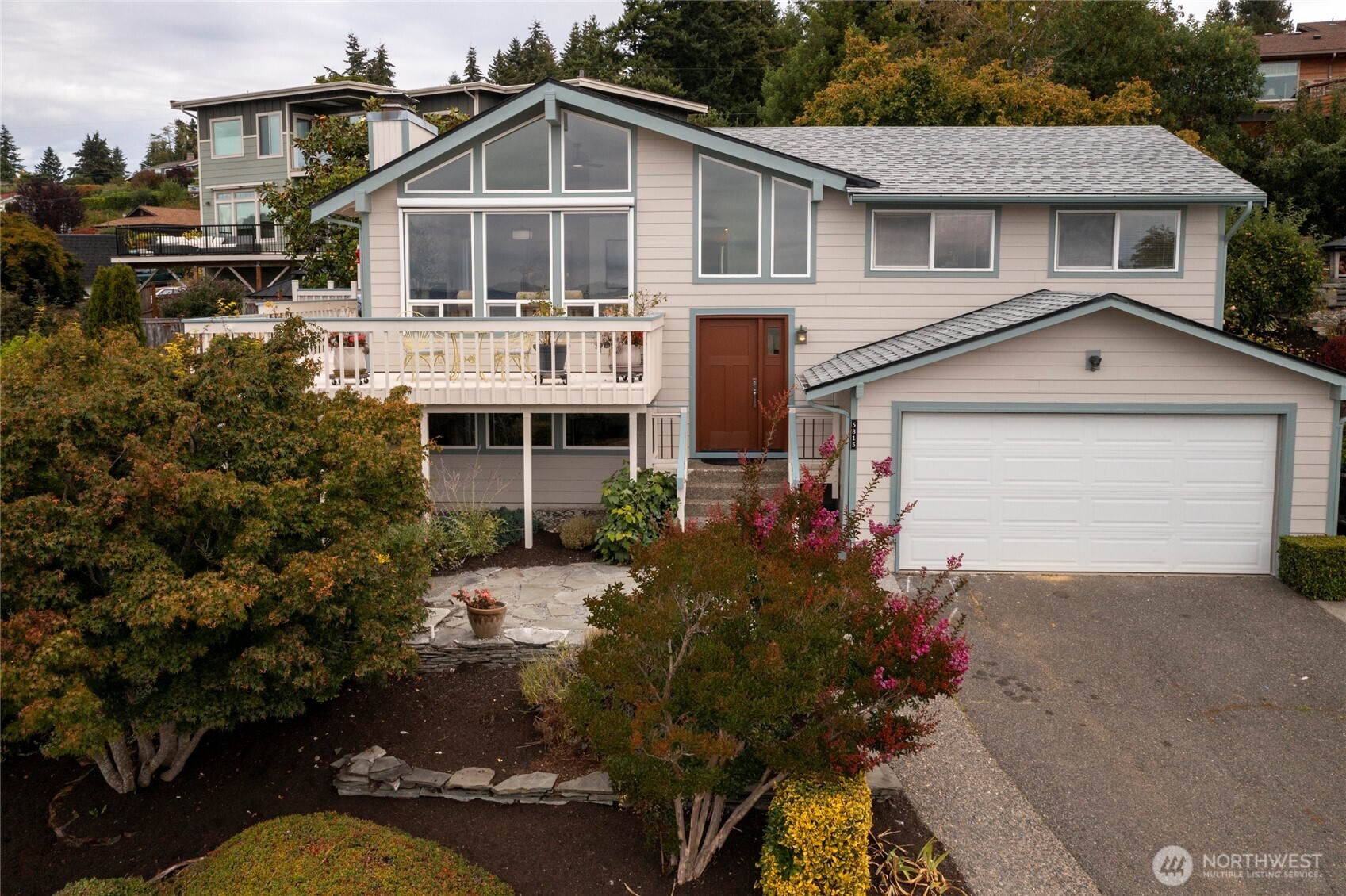 Gray split-level home with a white-railed deck, gabled windows, attached garage, and landscaped front garden.