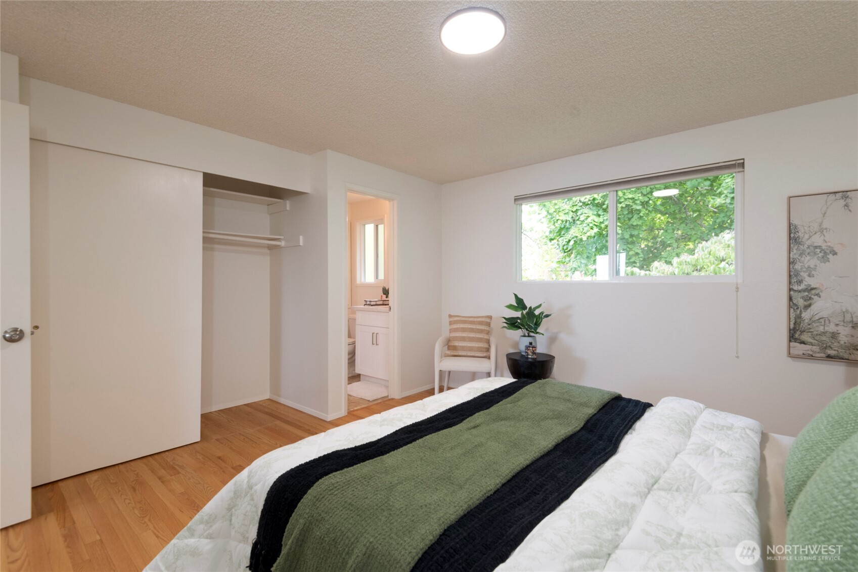 Staged bedroom with hardwood floors, an open closet, and a wide window overlooking greenery.