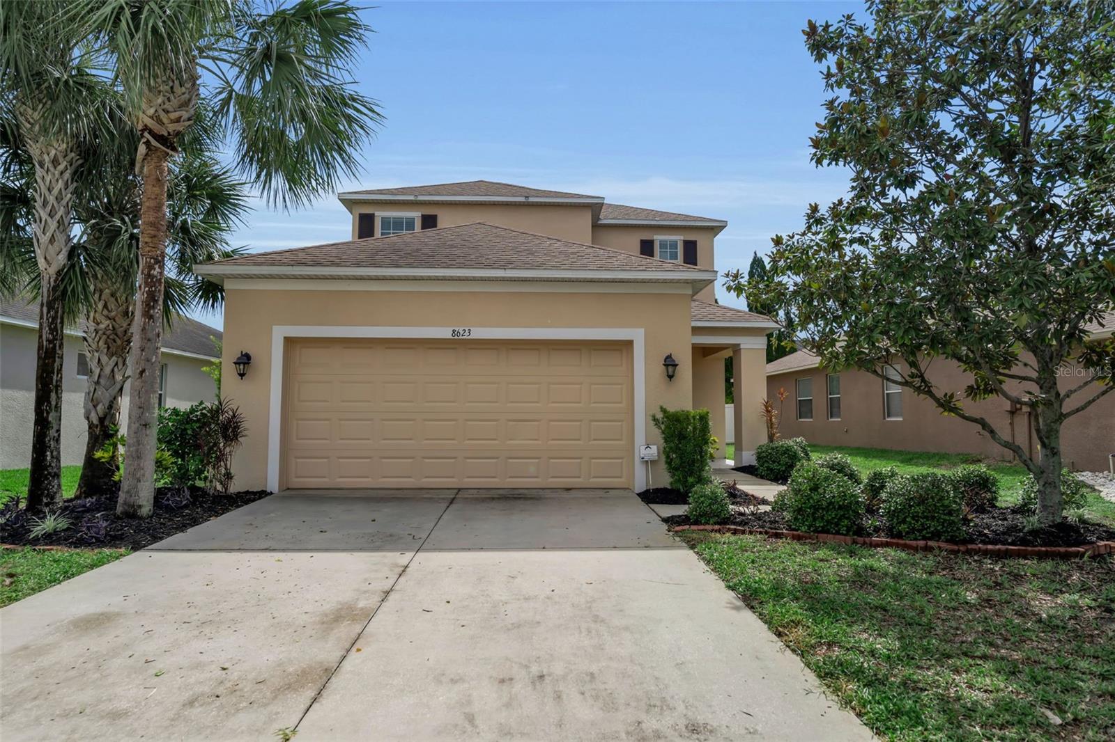Two-story beige house with a double garage, broad driveway, palm trees, and landscaped front yard.