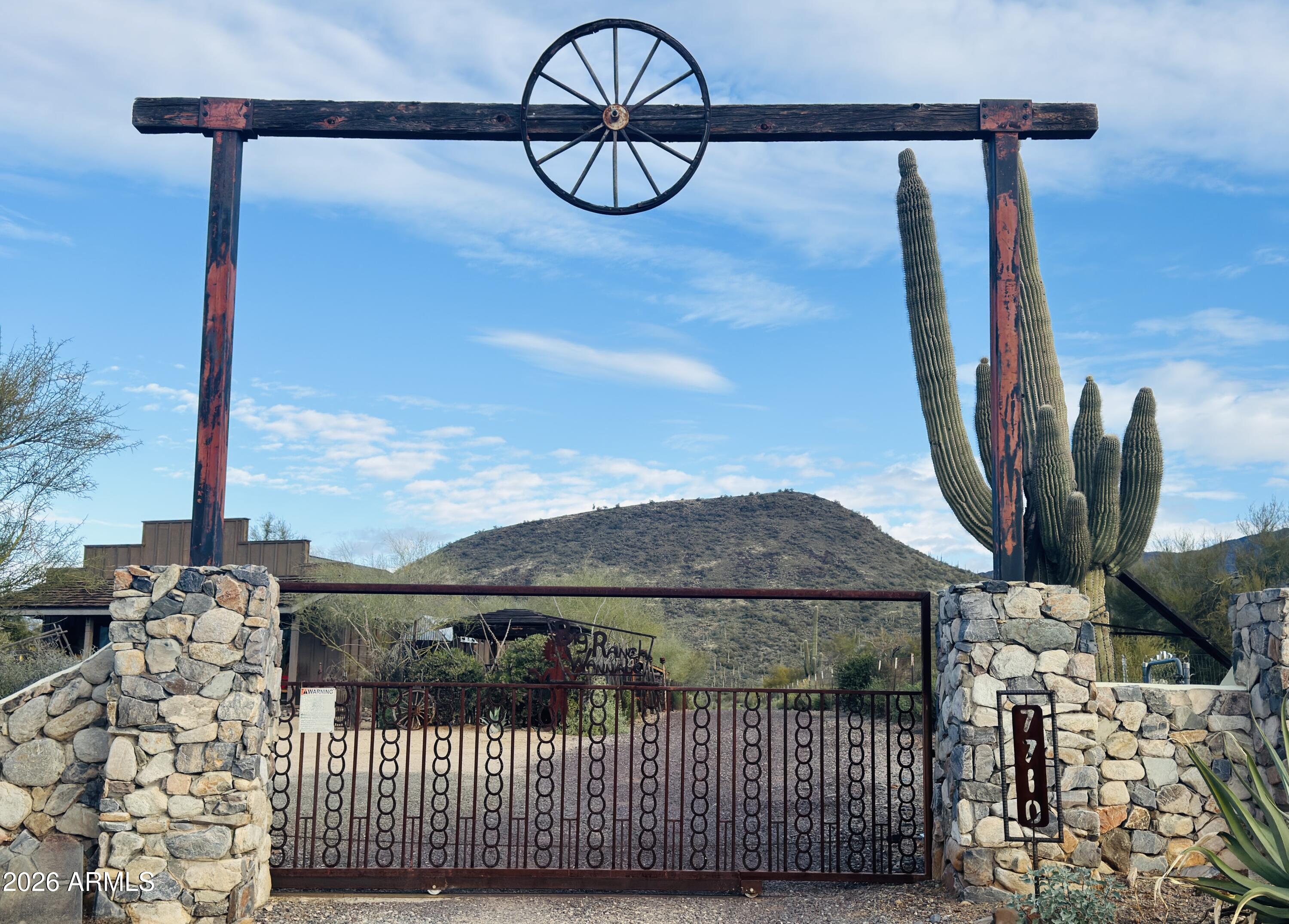 Rustic ranch gate framed by stone pillars, with saguaro cacti and a rocky desert hill.