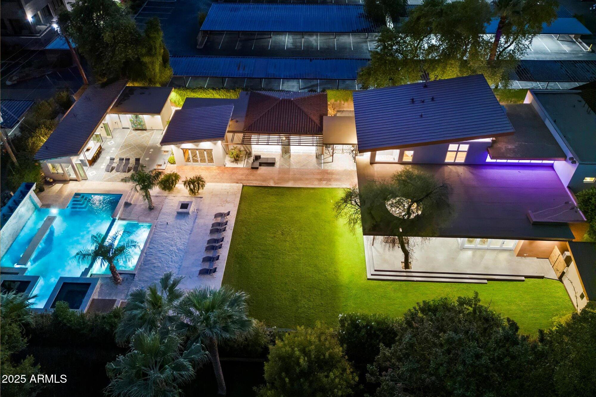 Aerial view of a modern home at night, with a bright swimming pool, landscaped lawn, and broad dark roofs.