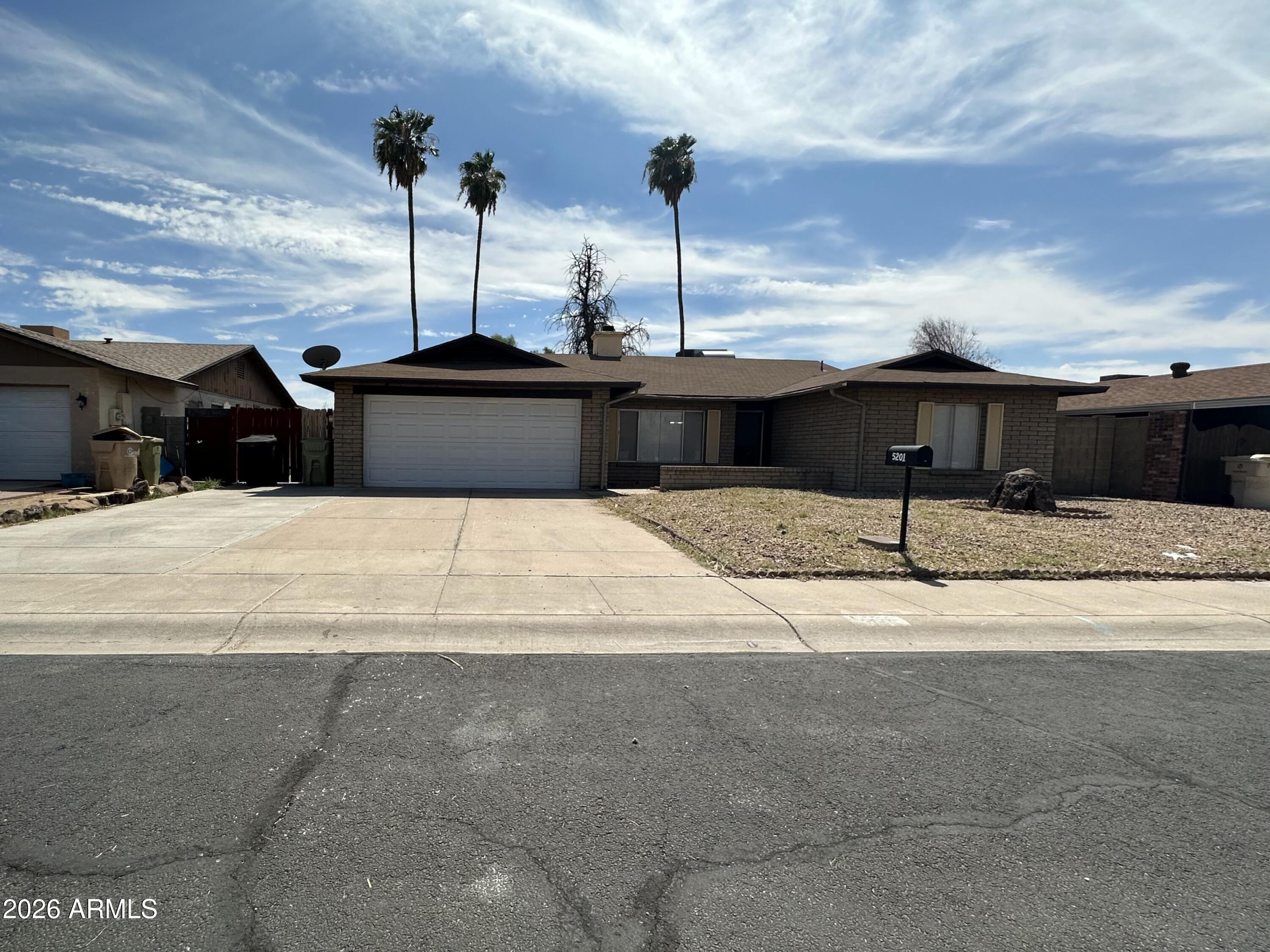 Single-story brick house with a two-car garage, broad driveway, and tall palm trees.