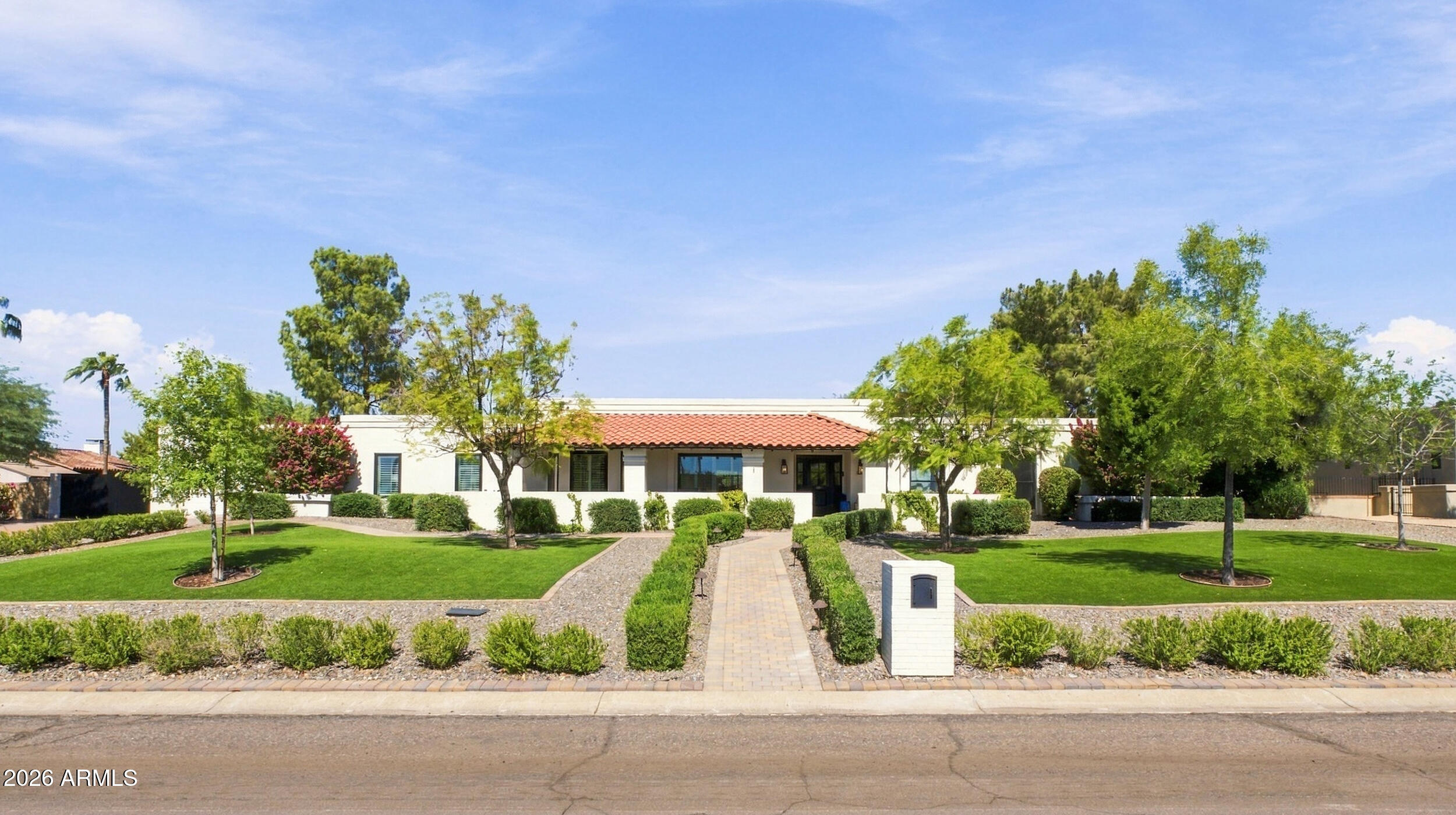 Single-story stucco home with a red tile roof, landscaped lawns, mature trees, and a central walkway.