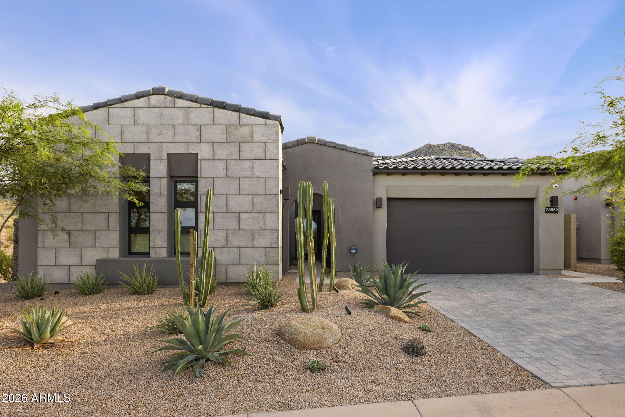 Modern desert home with stone facade, dark garage door, gravel landscaping, and mountain backdrop.