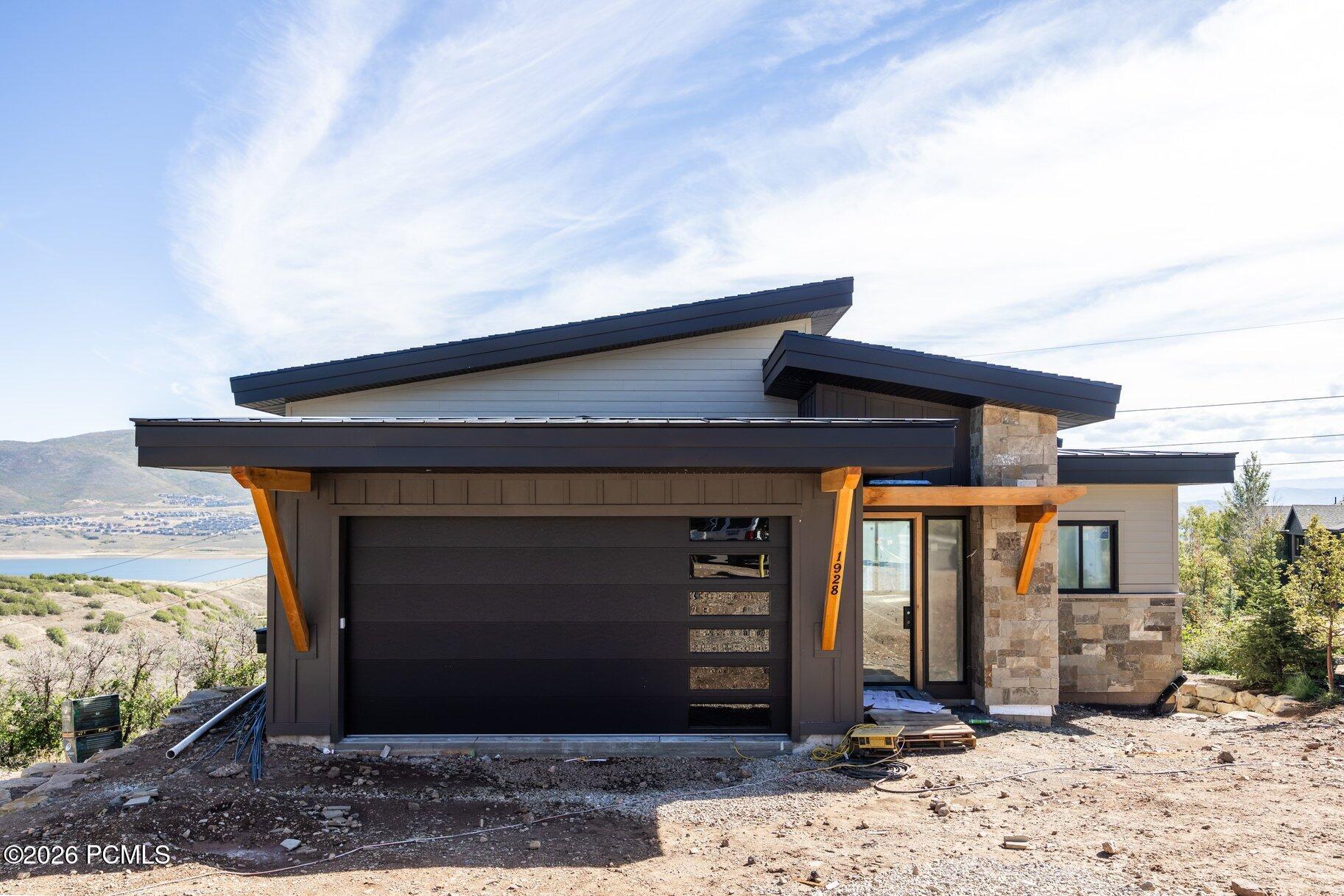 Modern home under construction with dramatic dark roofs, stone exterior, and a lake and hills behind it.