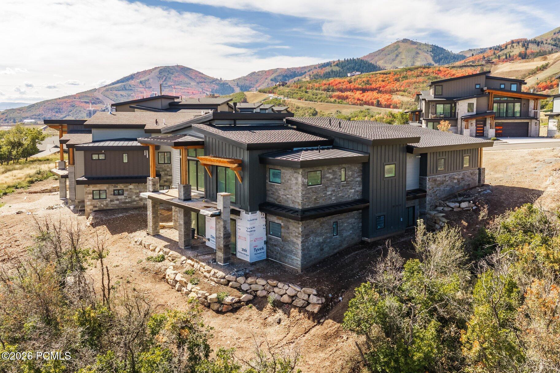 Modern home under construction on a hillside, with stone veneer, dark siding, and autumn mountains behind it.