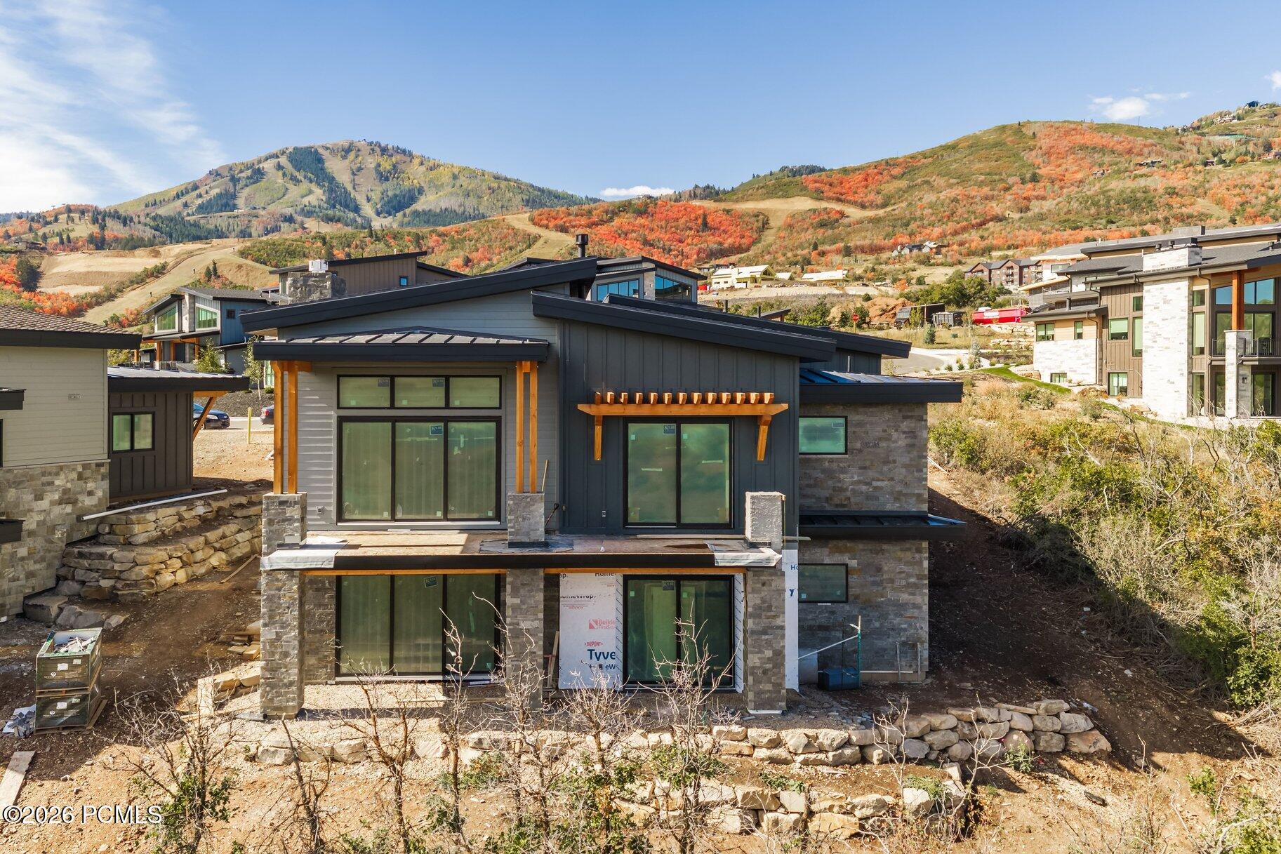 Modern mountain home under construction with dark siding, stonework, and autumn hills behind it.