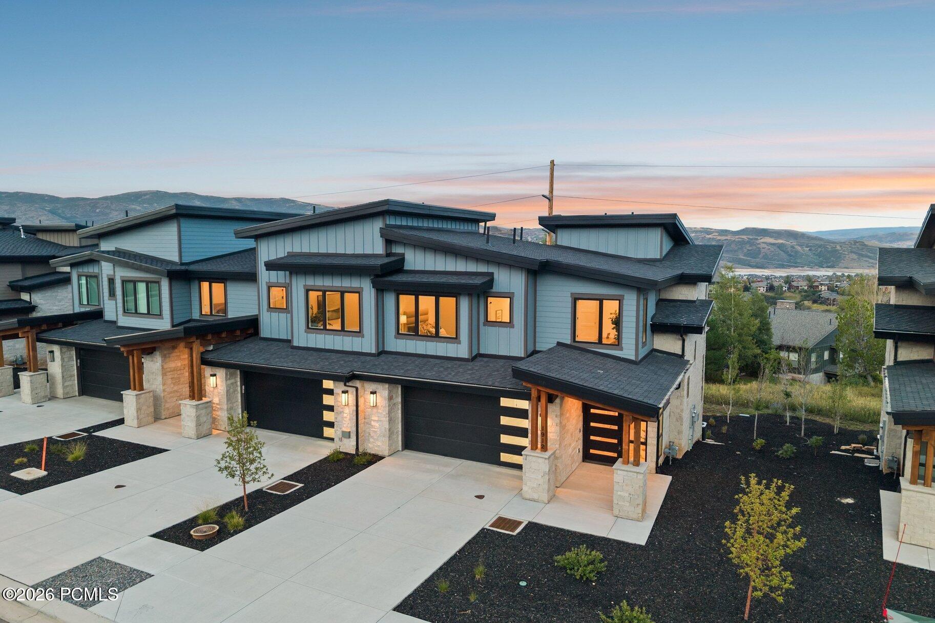 Modern blue-gray home with stone accents and black garage doors, lit warmly at dusk with mountains behind.