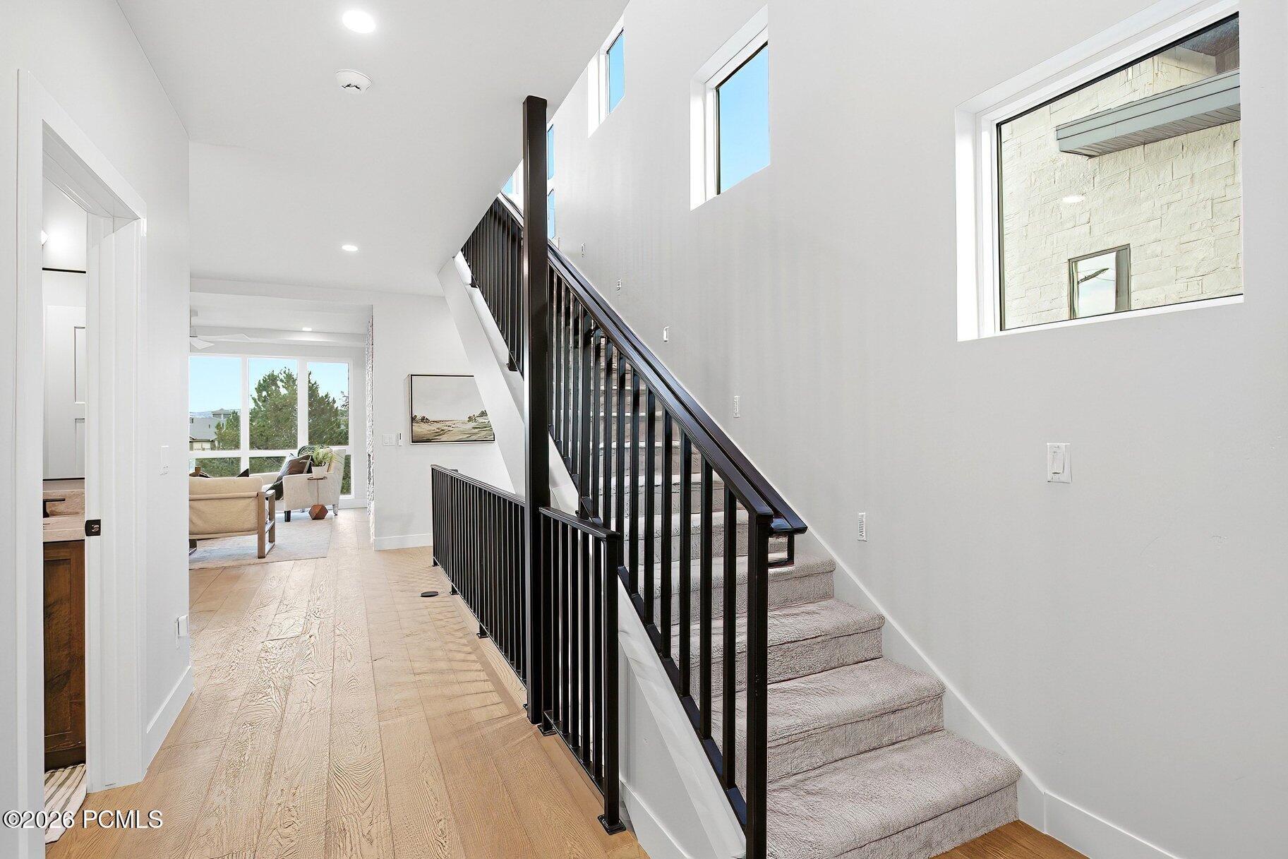 Bright entry hall with a carpeted staircase, black railings, and a windowed sitting area beyond.