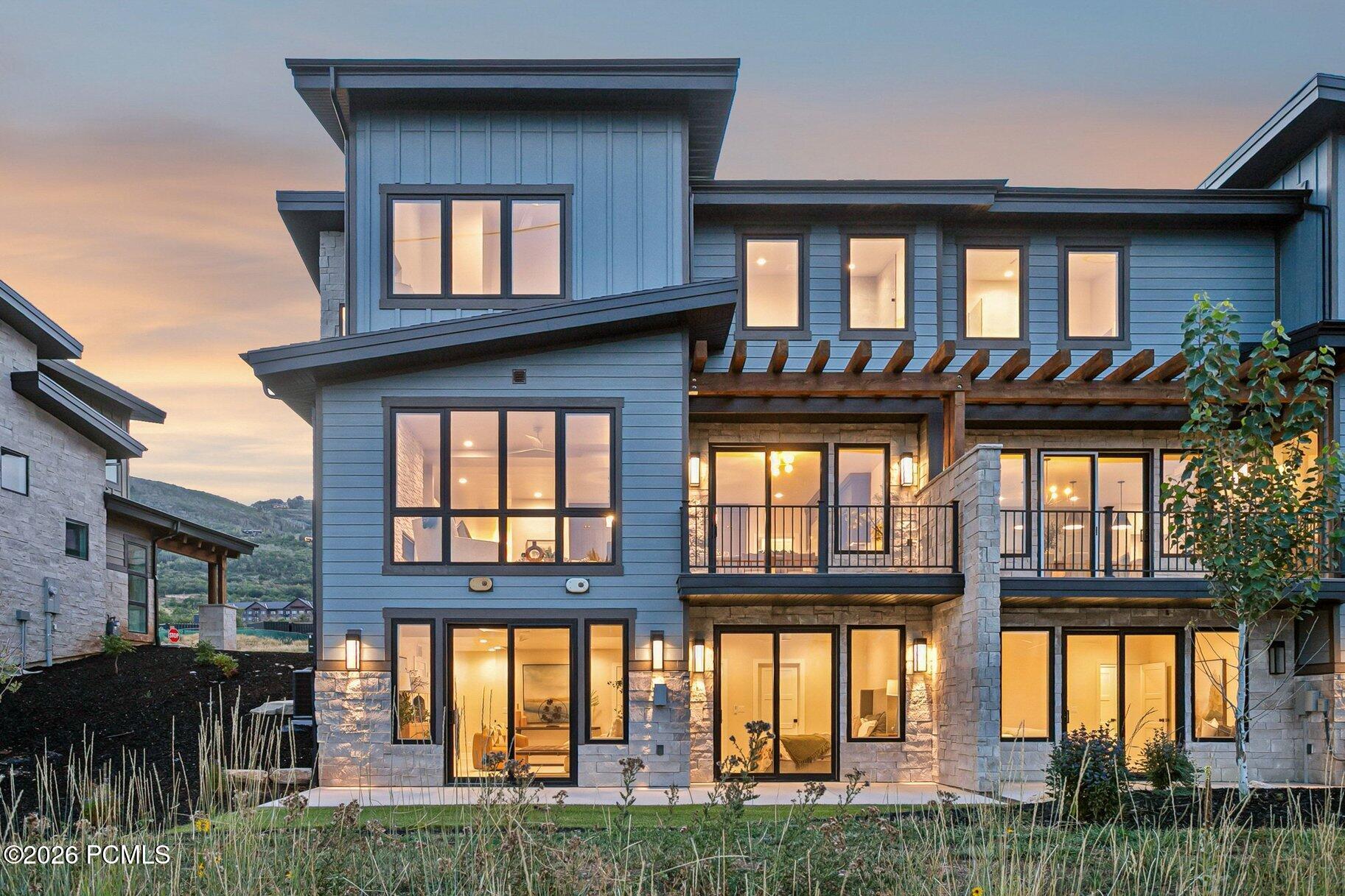 Modern blue-gray home with stone accents and a timber pergola, warmly lit at dusk.