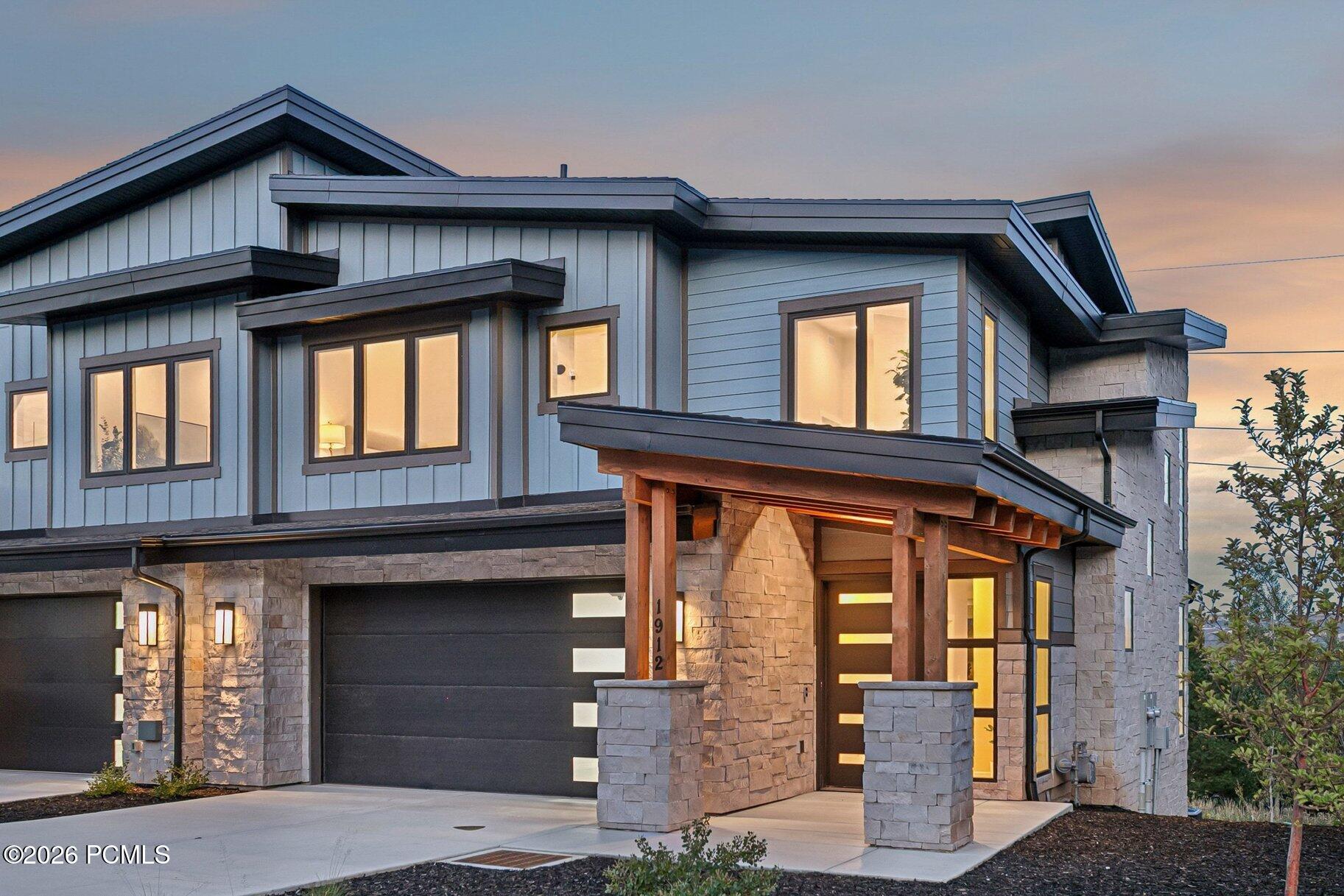 Modern two-story home with stone walls, dark garage doors, and a warmly lit covered entry at dusk.