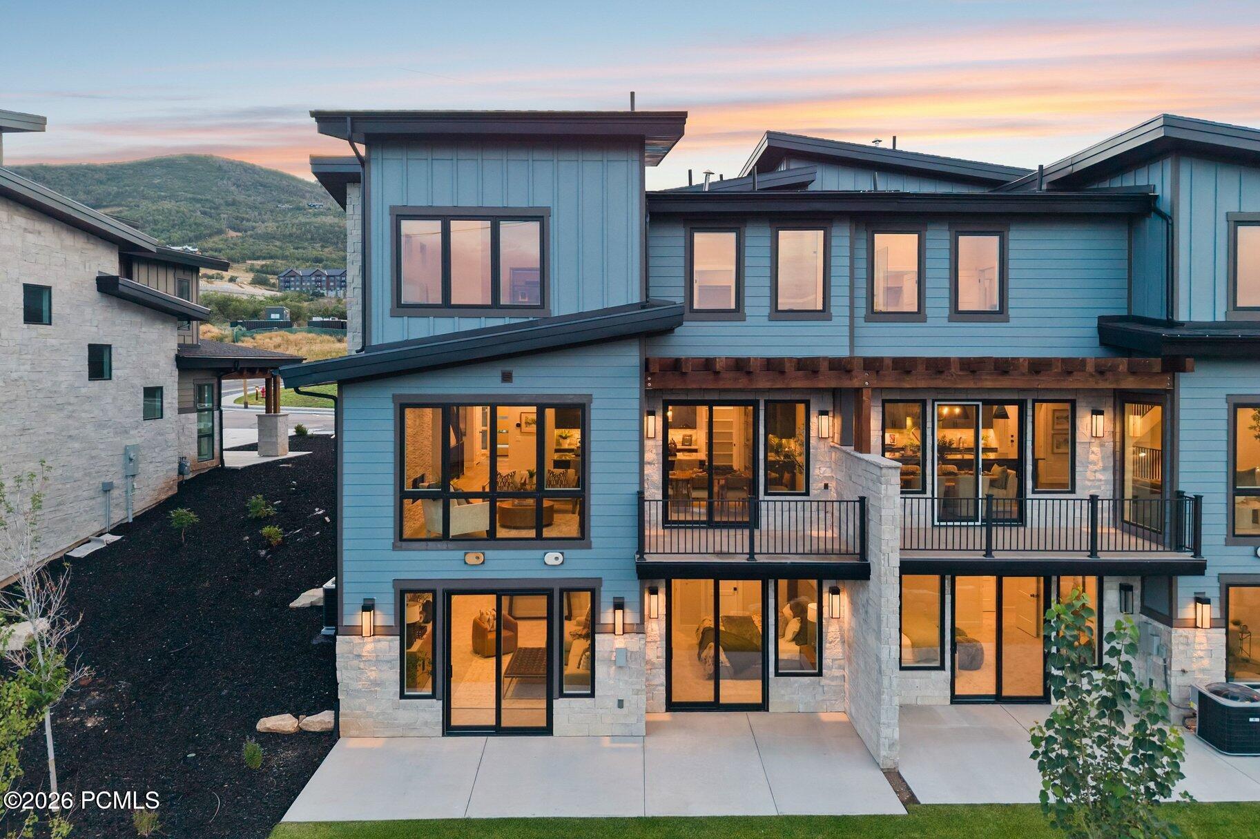 Modern blue multistory home with stone accents and balconies, warmly lit at dusk against a mountain hillside.