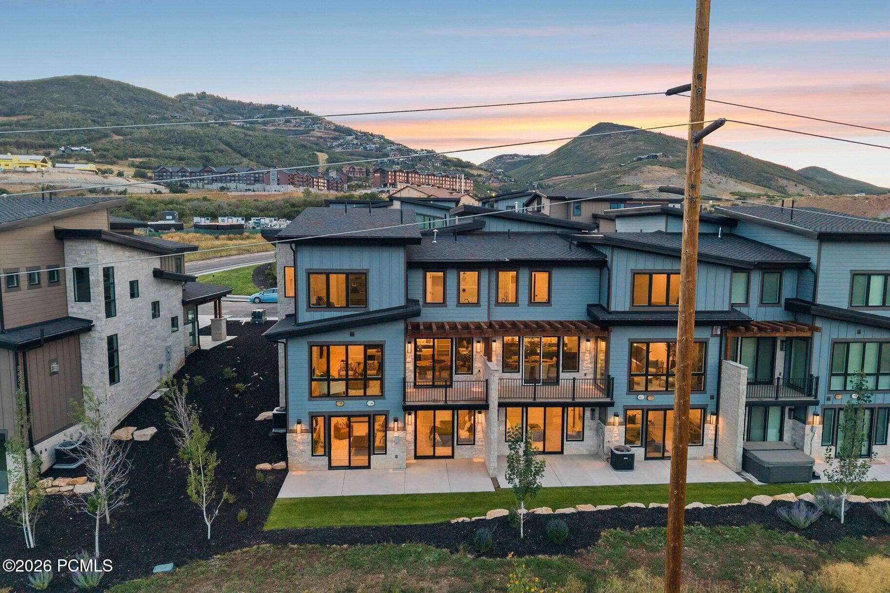 Modern blue-gray home with stone accents and balconies, warmly lit at dusk against hillside homes.
