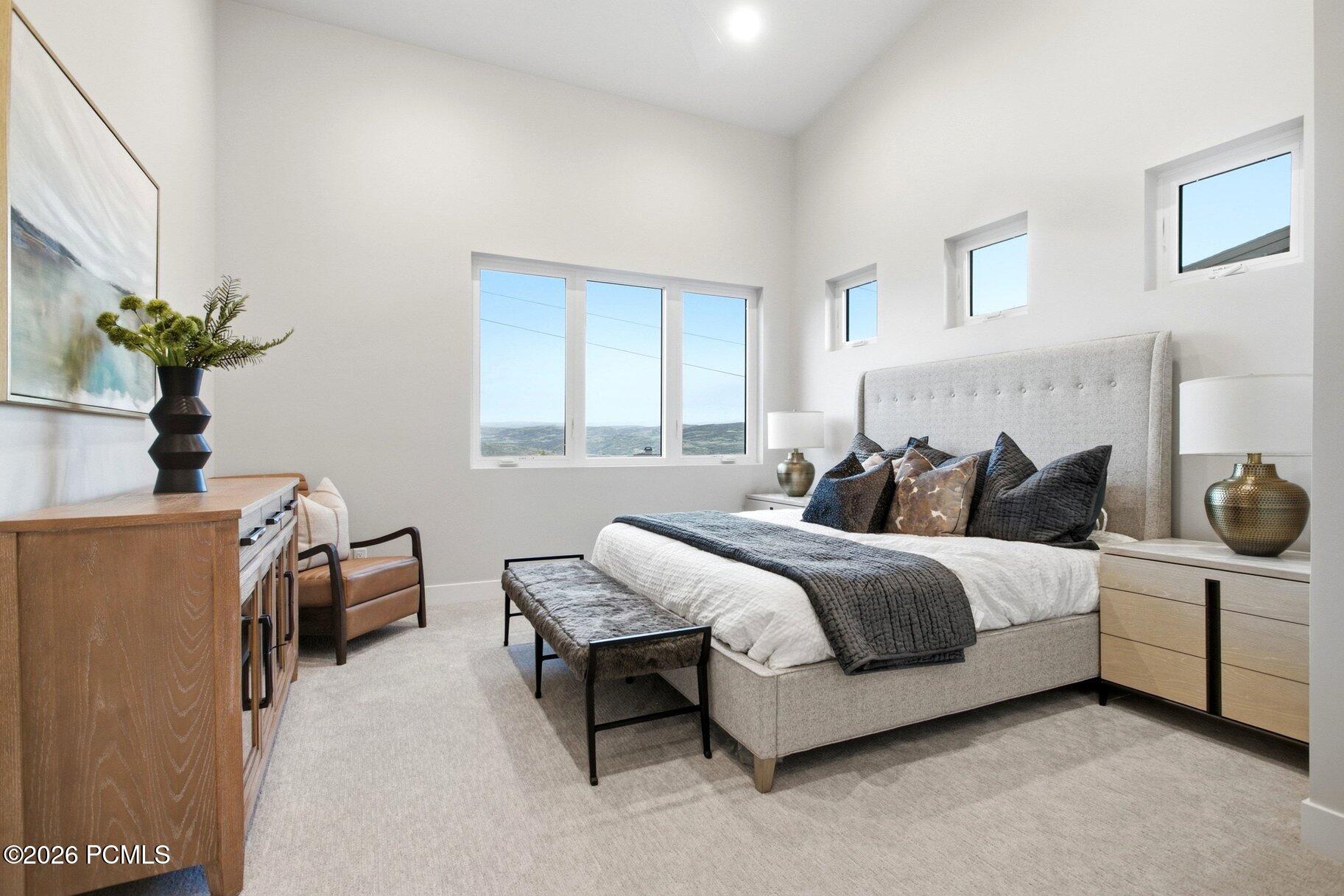 Staged bedroom with a gray upholstered bed and broad windows overlooking distant hills.
