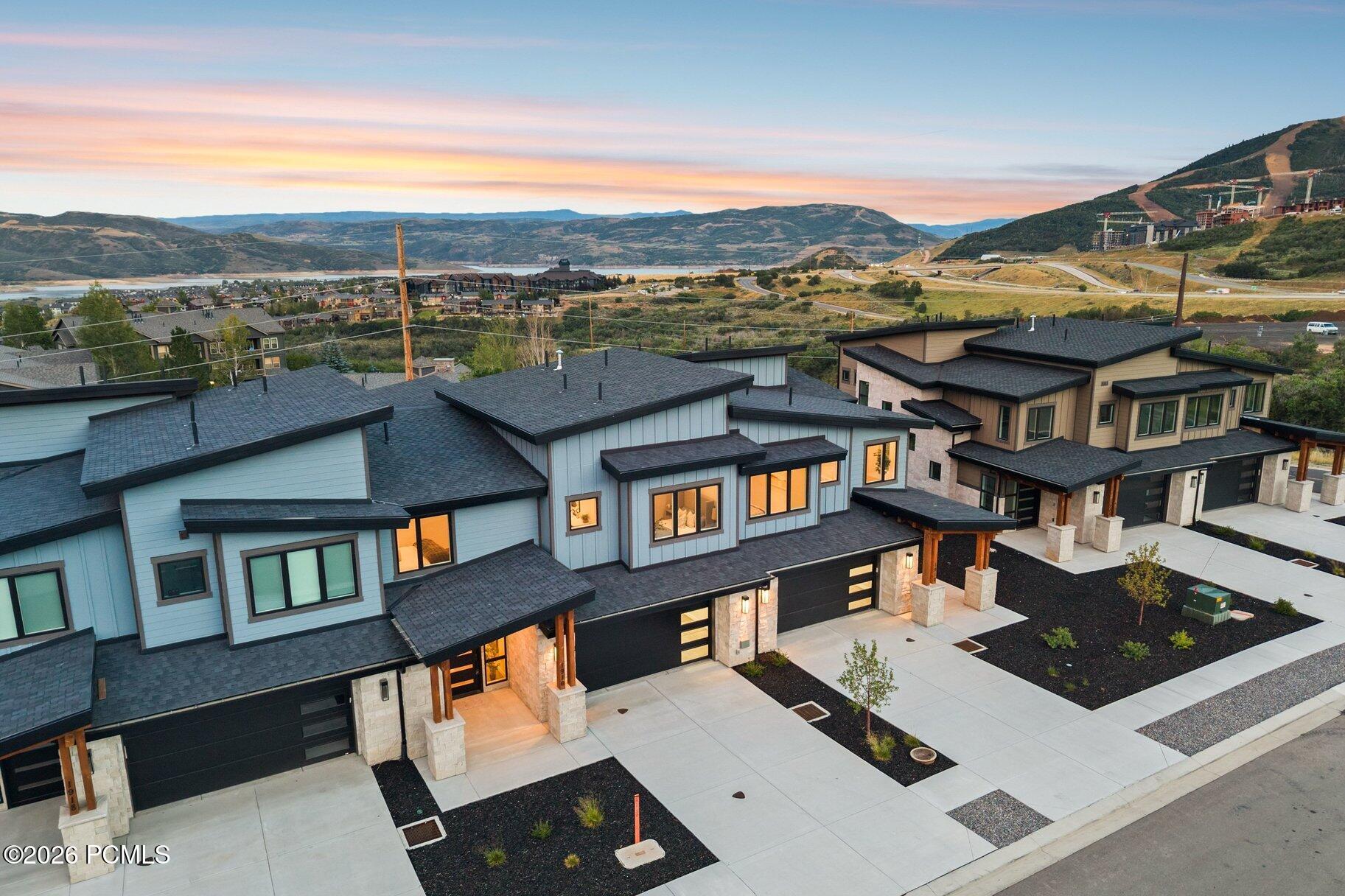 Modern homes with illuminated windows overlook a lake and mountain valley at sunset.