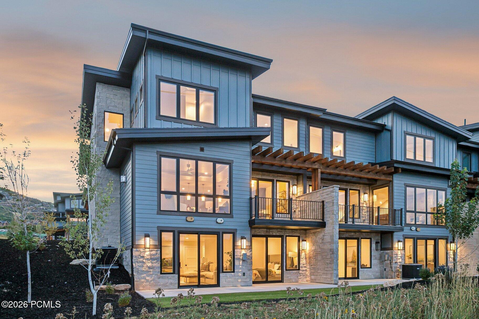 Modern blue-gray home with stone accents, illuminated windows, balconies, and a timber pergola at dusk.