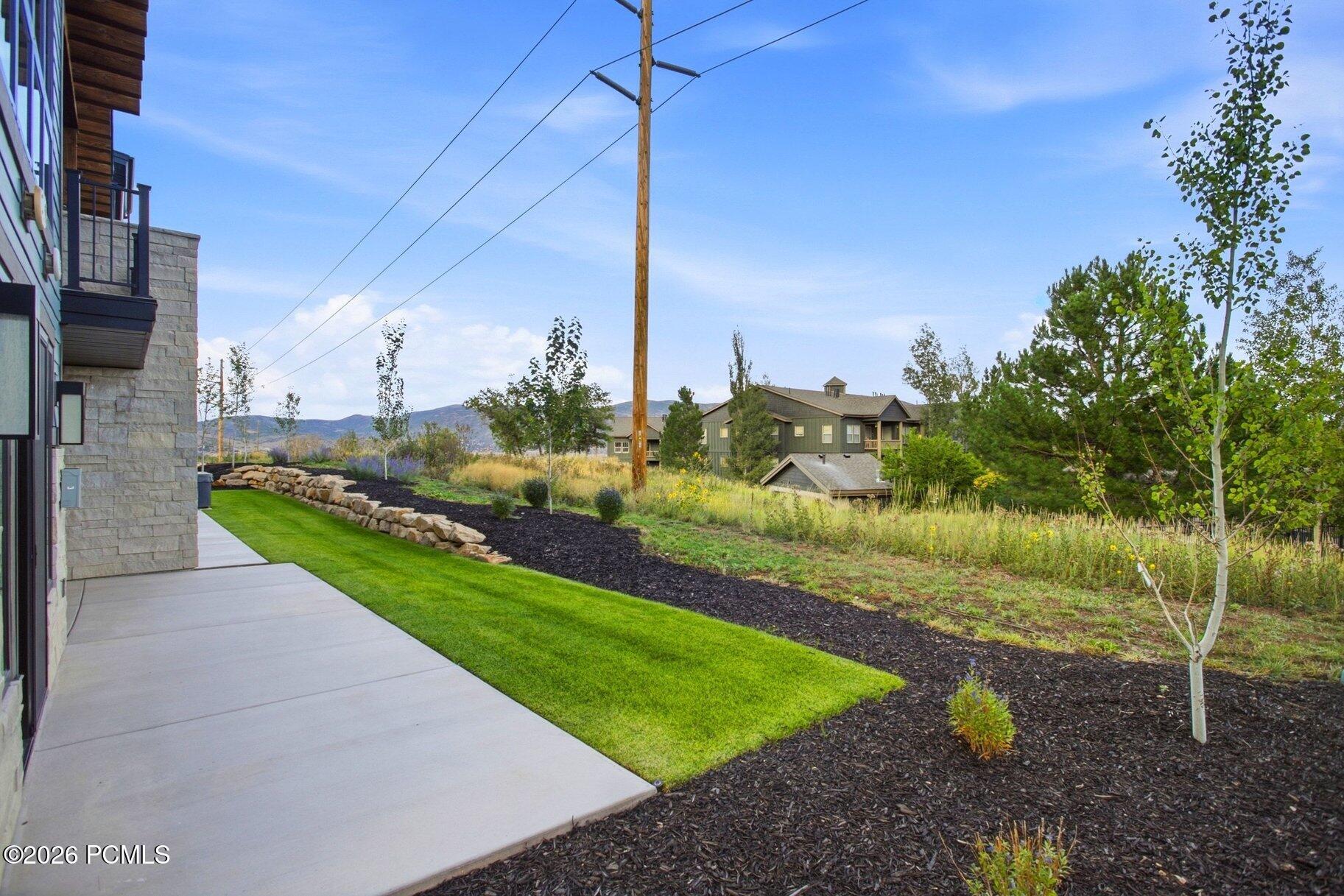 Side yard with a concrete walkway, green lawn, landscaped beds, young trees, and distant hills.