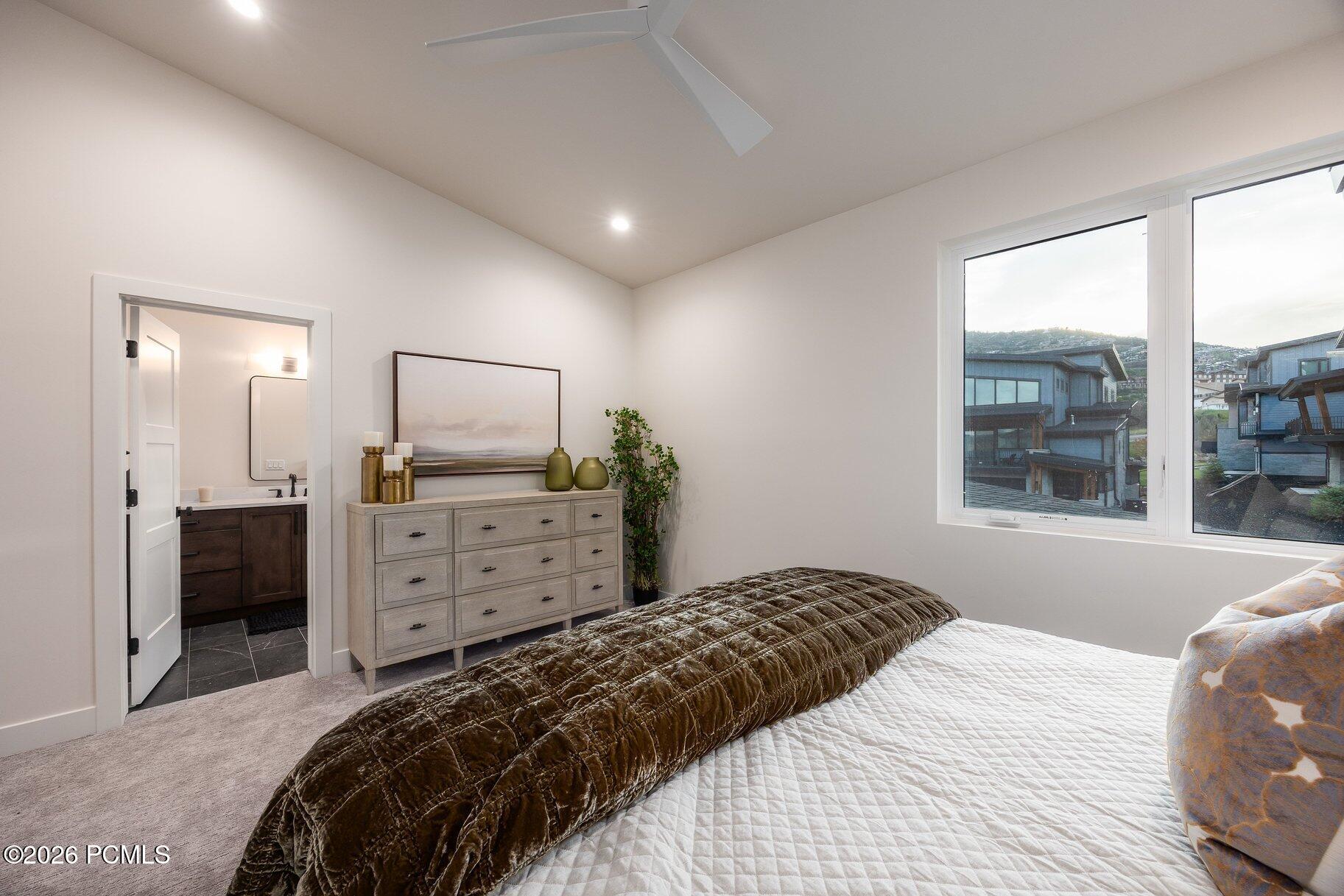 Bedroom with a dark velvet coverlet, light wood dresser, and large windows overlooking neighboring homes.