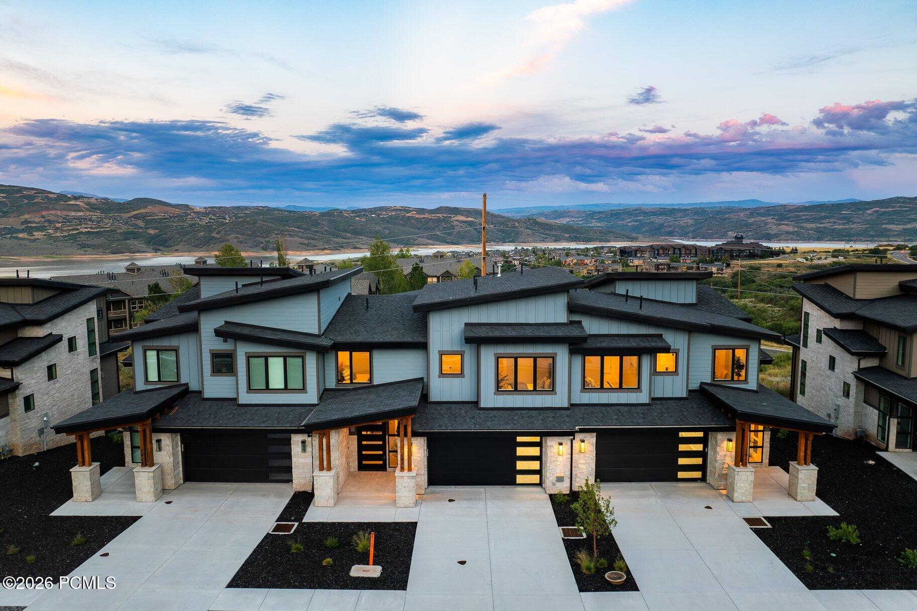 Modern home with glowing windows overlooks a lake and rolling hills at dusk.