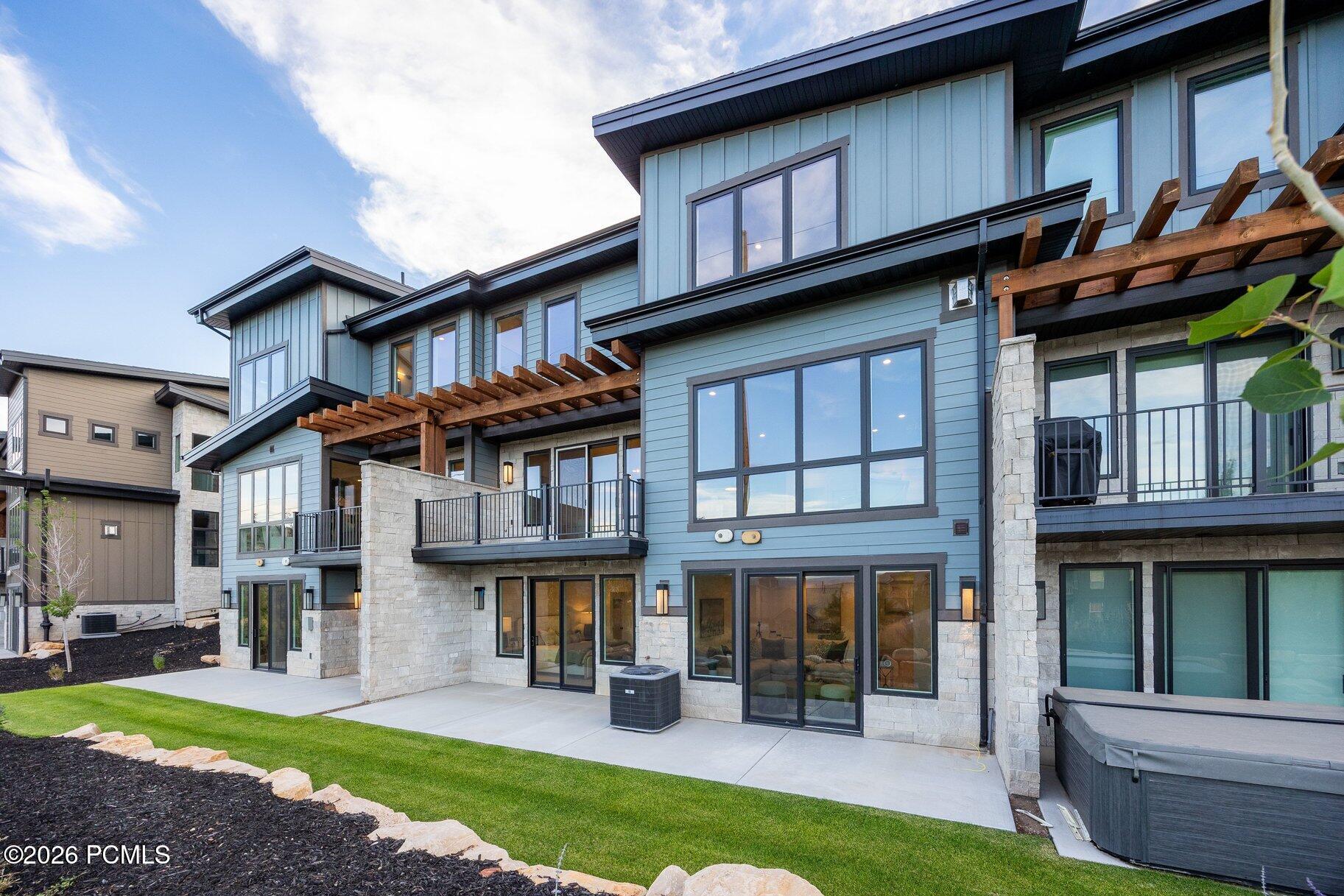 Modern blue-gray home with stone accents, balconies, wooden pergolas, landscaped lawn, and a covered hot tub.