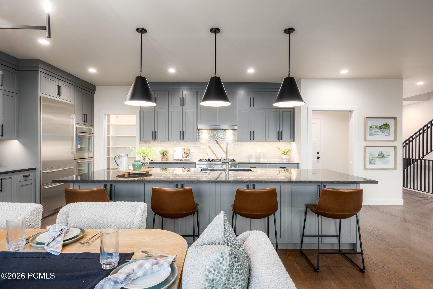 Staged kitchen with gray cabinets, a large island, stainless appliances, and three pendant lights.