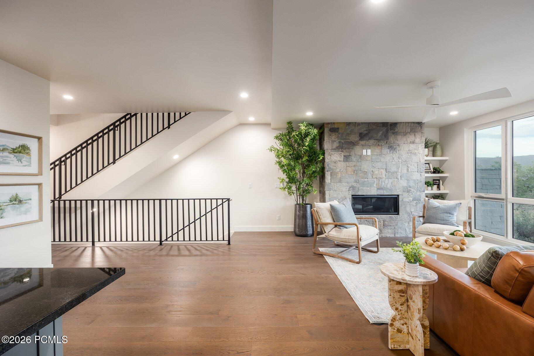 Staged living room with a stone fireplace, black-railed staircase, wood floors, and large windows overlooking hills.