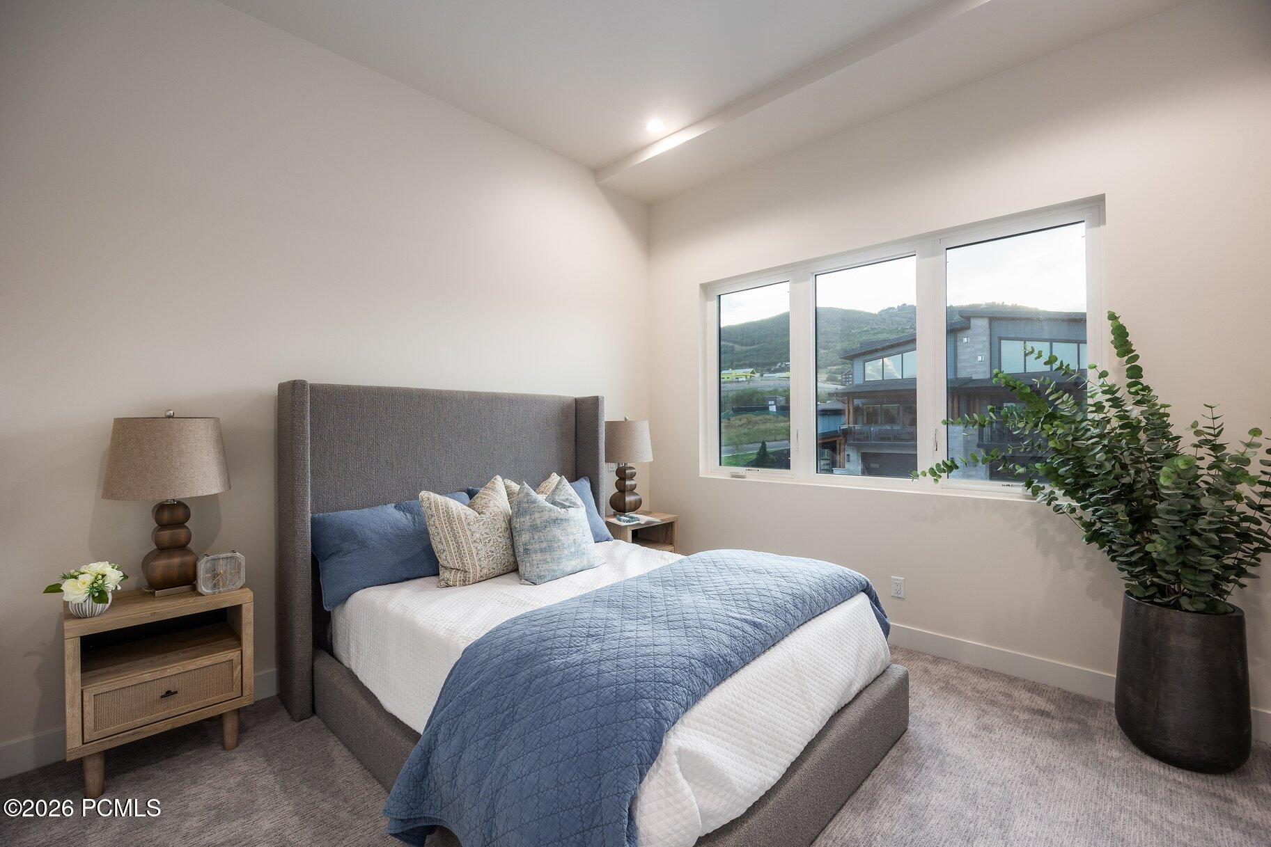 Staged bedroom with a gray upholstered bed and large window overlooking green hills.