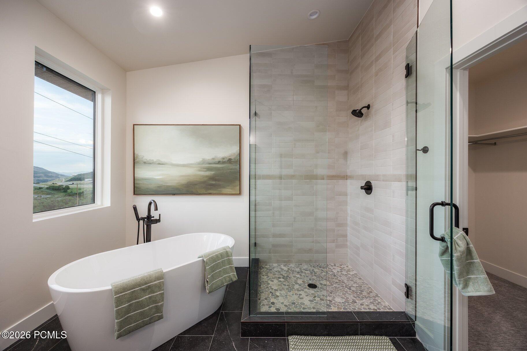 Modern bathroom with a freestanding soaking tub, glass walk-in shower, and window overlooking green hills.