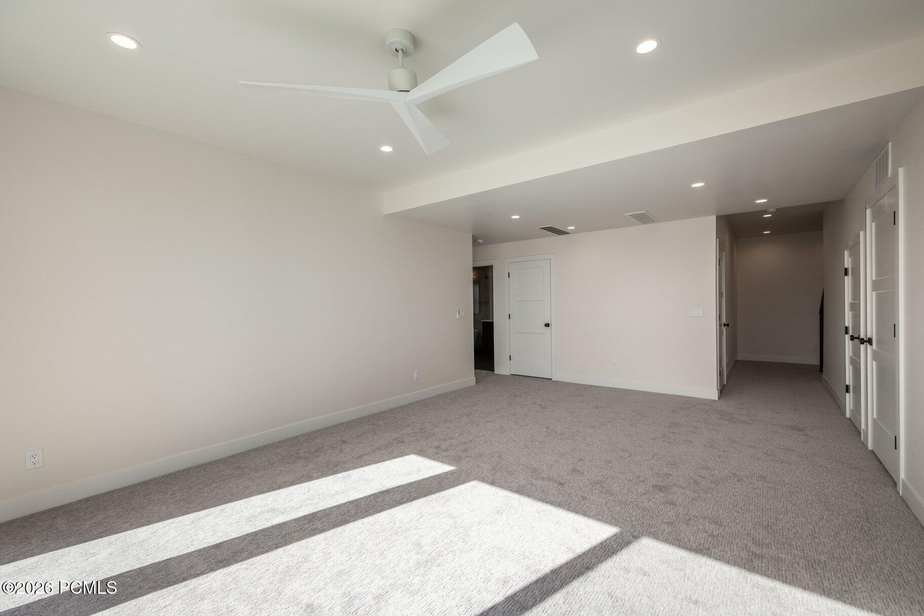 Empty carpeted room with a ceiling fan, several doors, and bright sunlight across the floor.