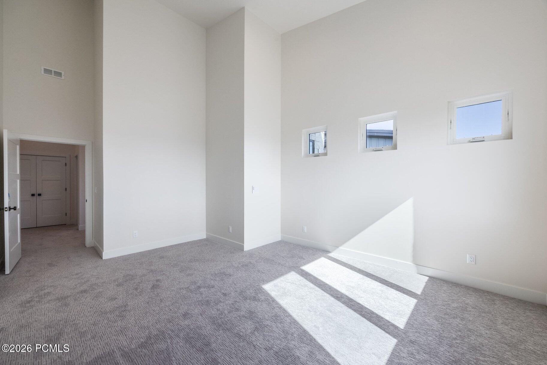 Empty carpeted room with a high ceiling and sunlight streaming through three small windows.