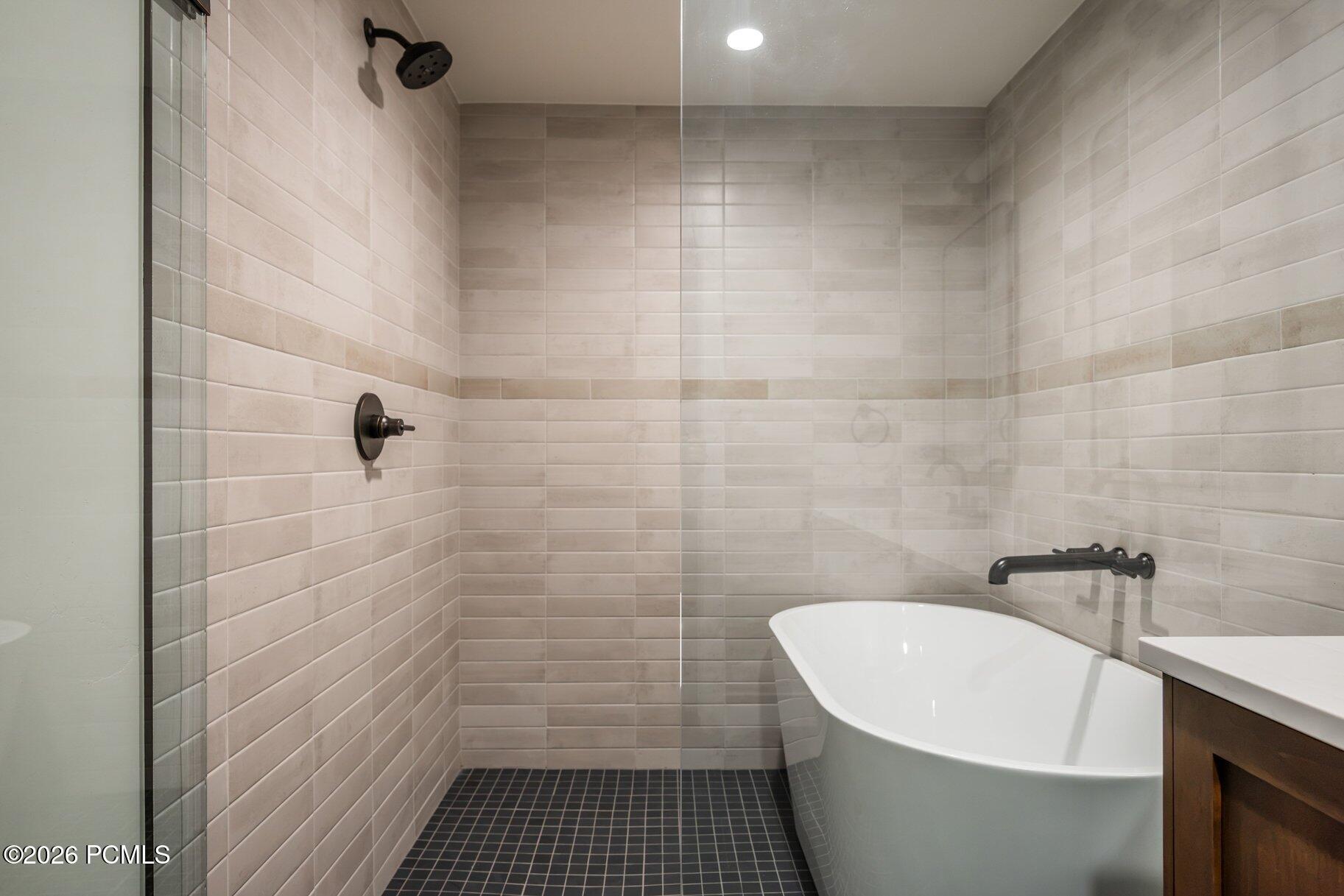 Modern bathroom with a freestanding tub, glass shower, pale tile walls, and dark fixtures.