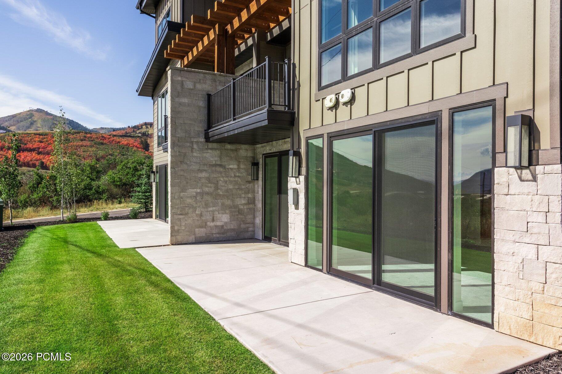 Modern home exterior with a stone facade, glass patio doors, balcony, and autumn hills beyond the lawn.