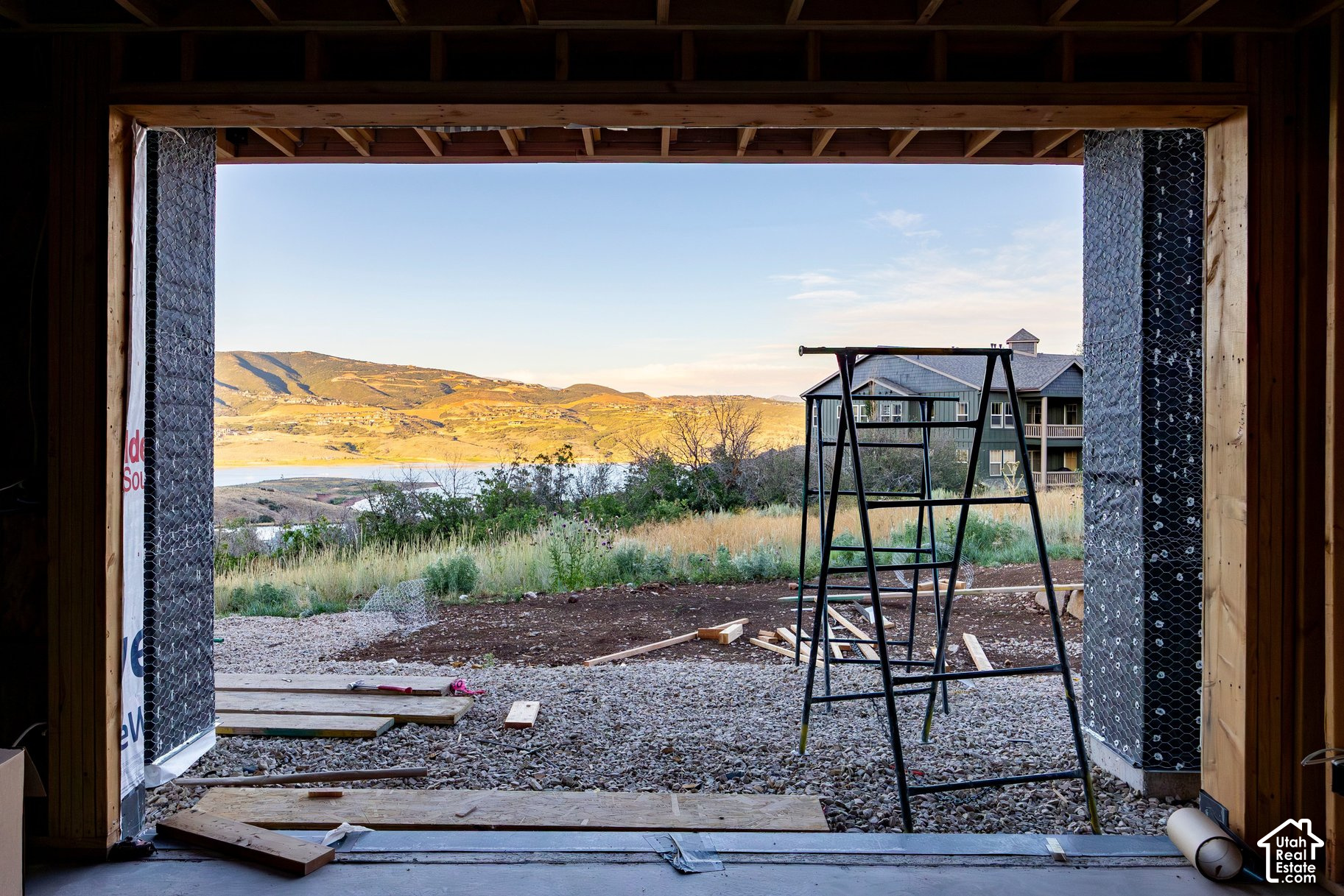 Unfinished structure opens onto a lake and golden hills, with a neighboring home beyond the construction area.