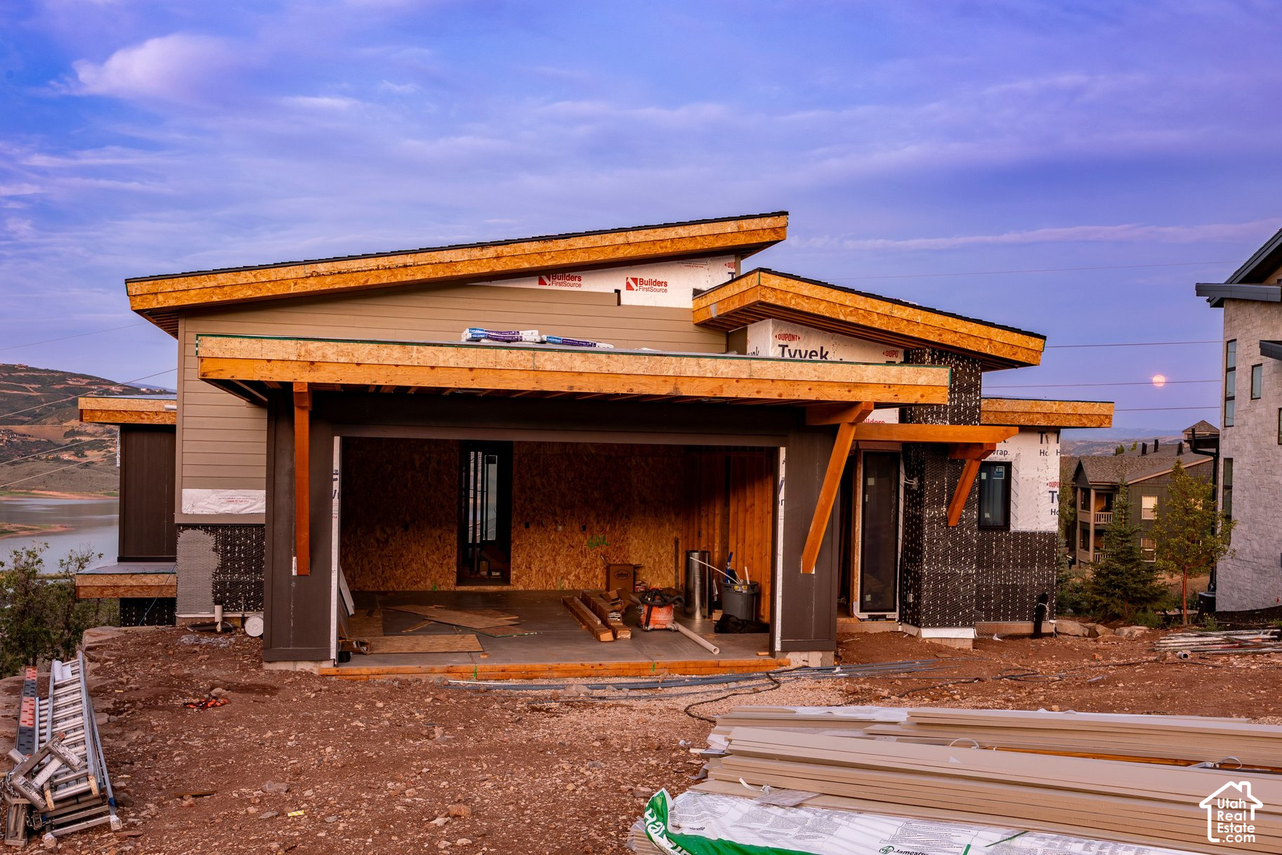 Modern home under construction with angular rooflines, overlooking a lake at dusk.